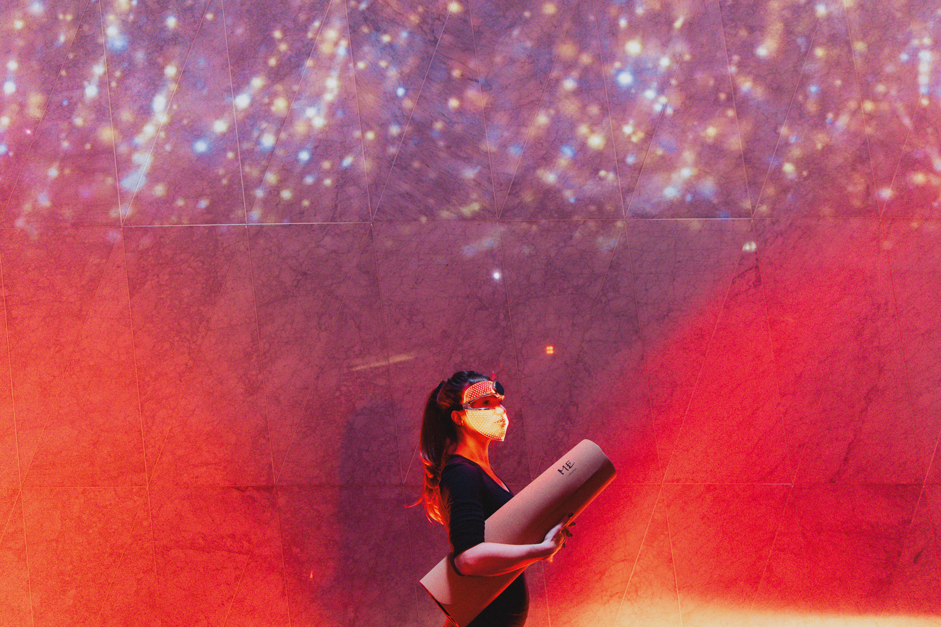 Woman in glowing LED mask holding yoga mat against sparkling cosmic wall.