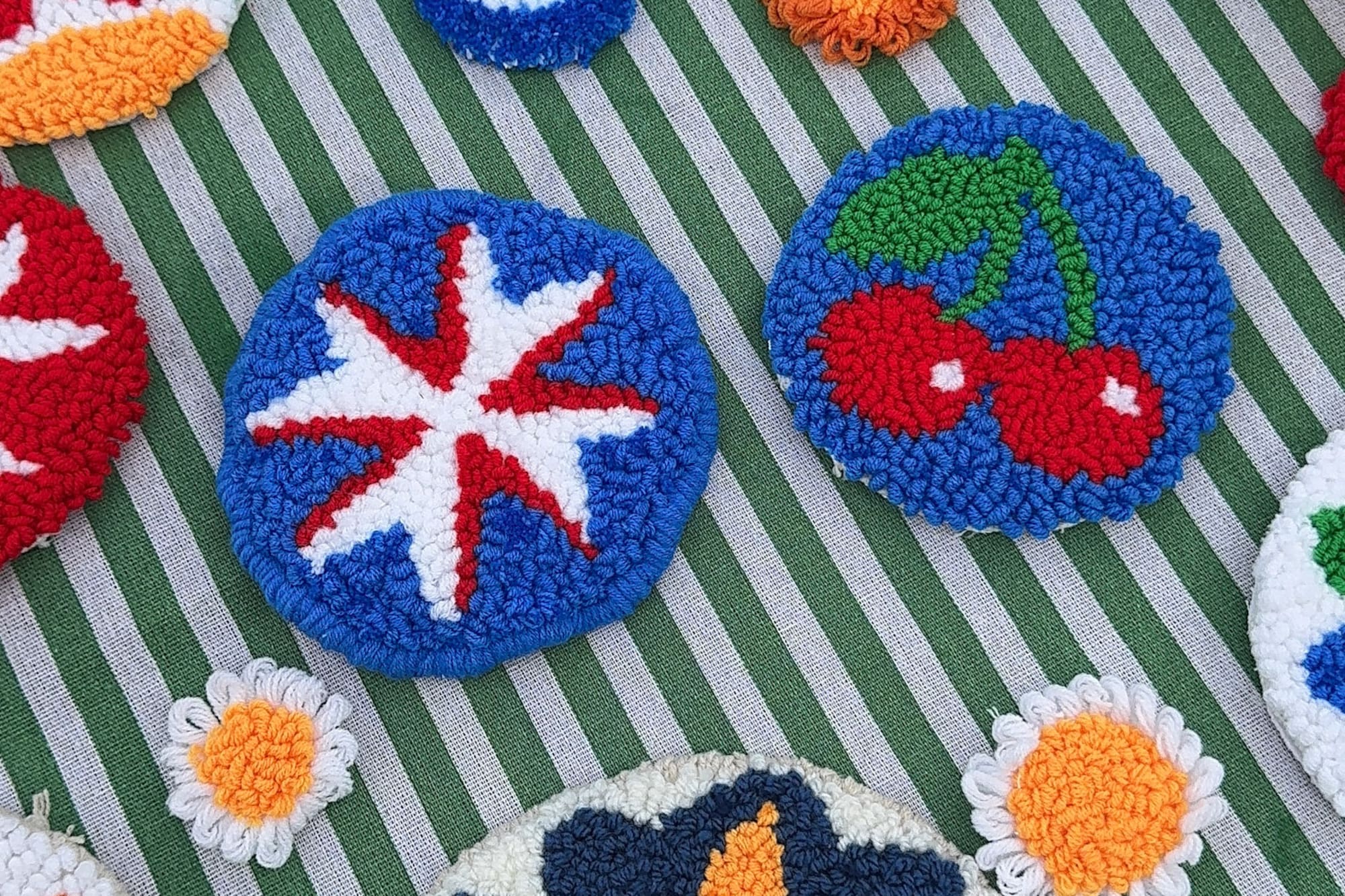 Colorful punch needle coasters with diverse patterns on green striped fabric.