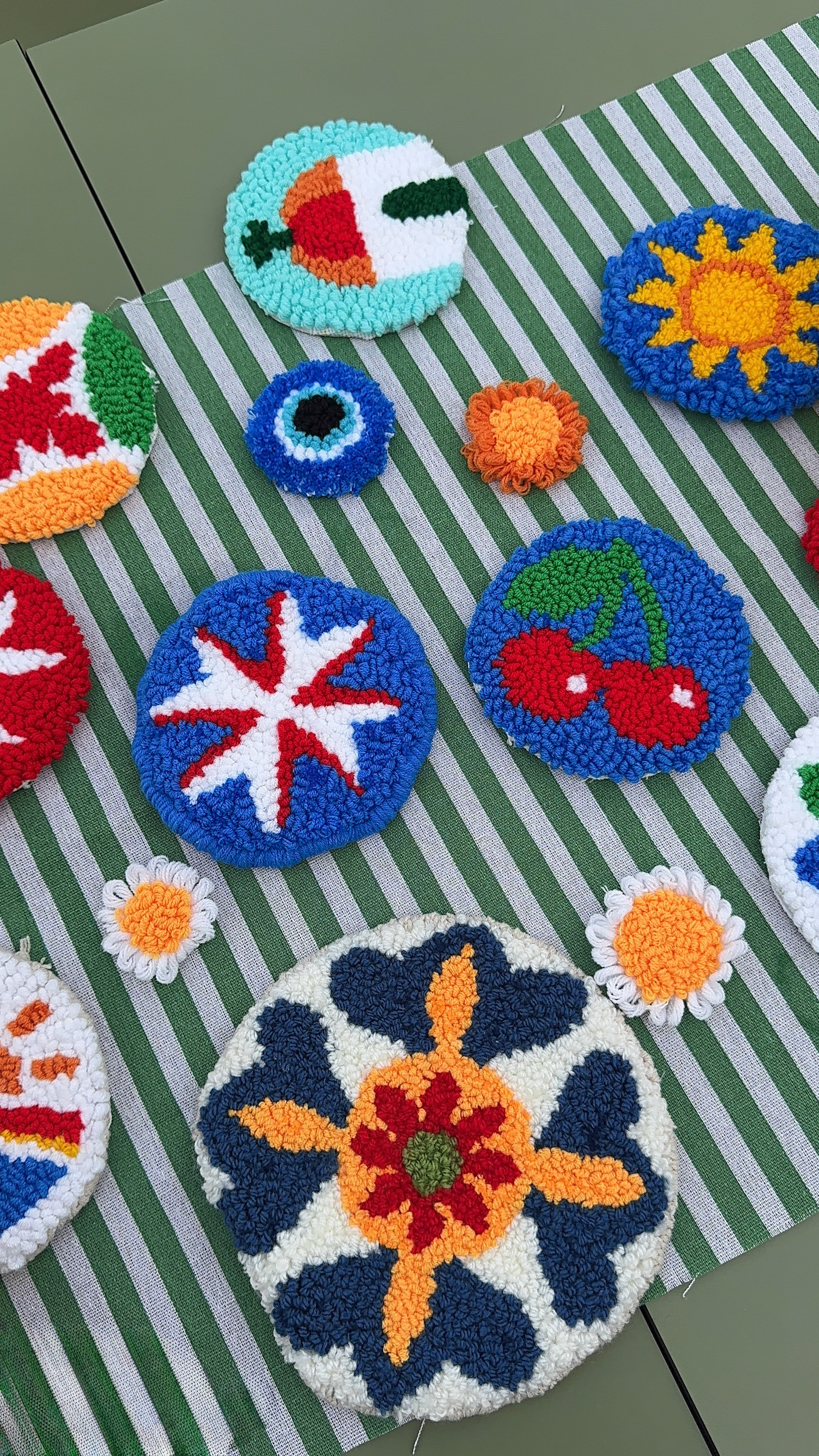 Colorful punch needle coasters with diverse patterns on green striped fabric.