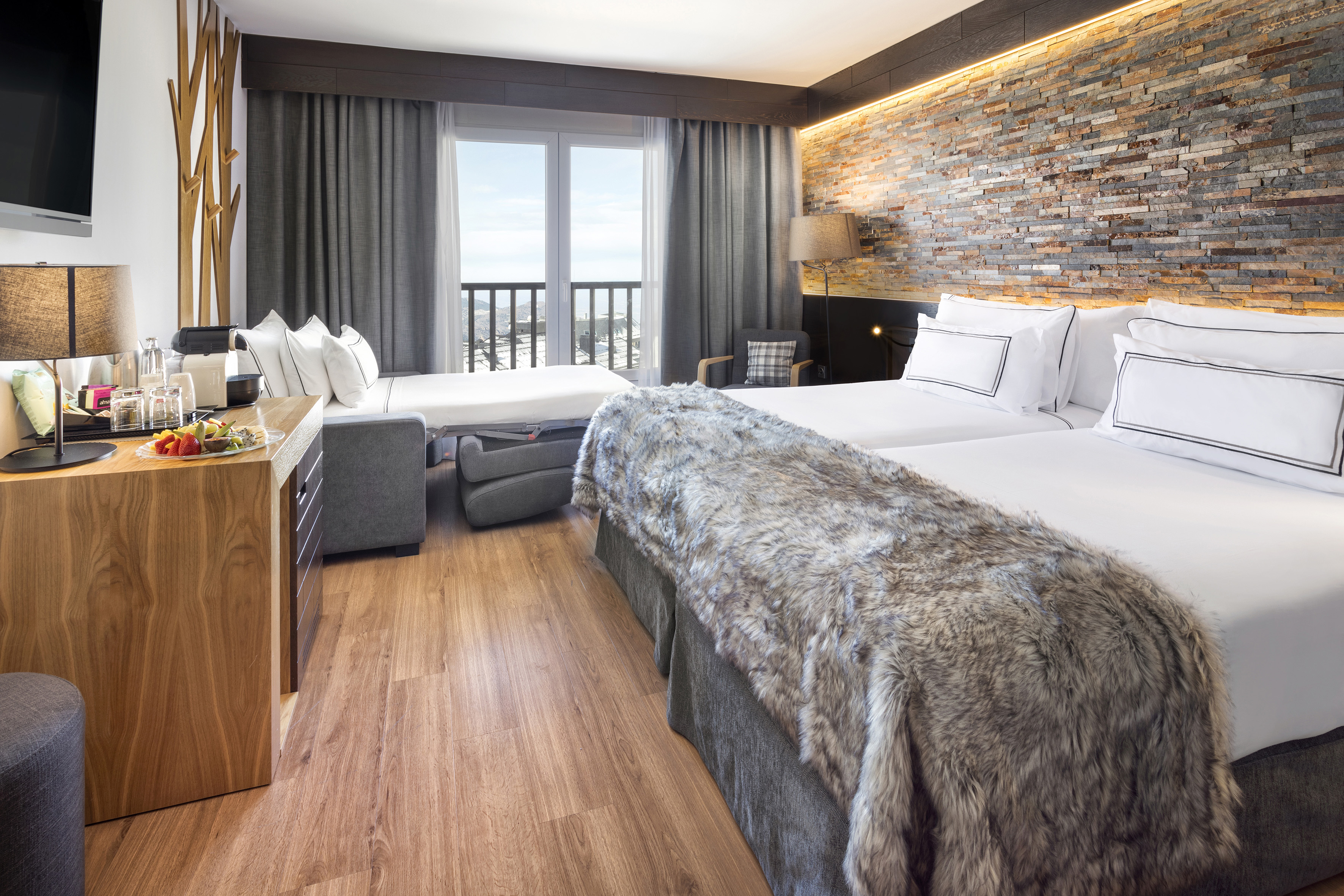 Mountain hotel room with twin beds, sofa bed, stone wall, and balcony view.