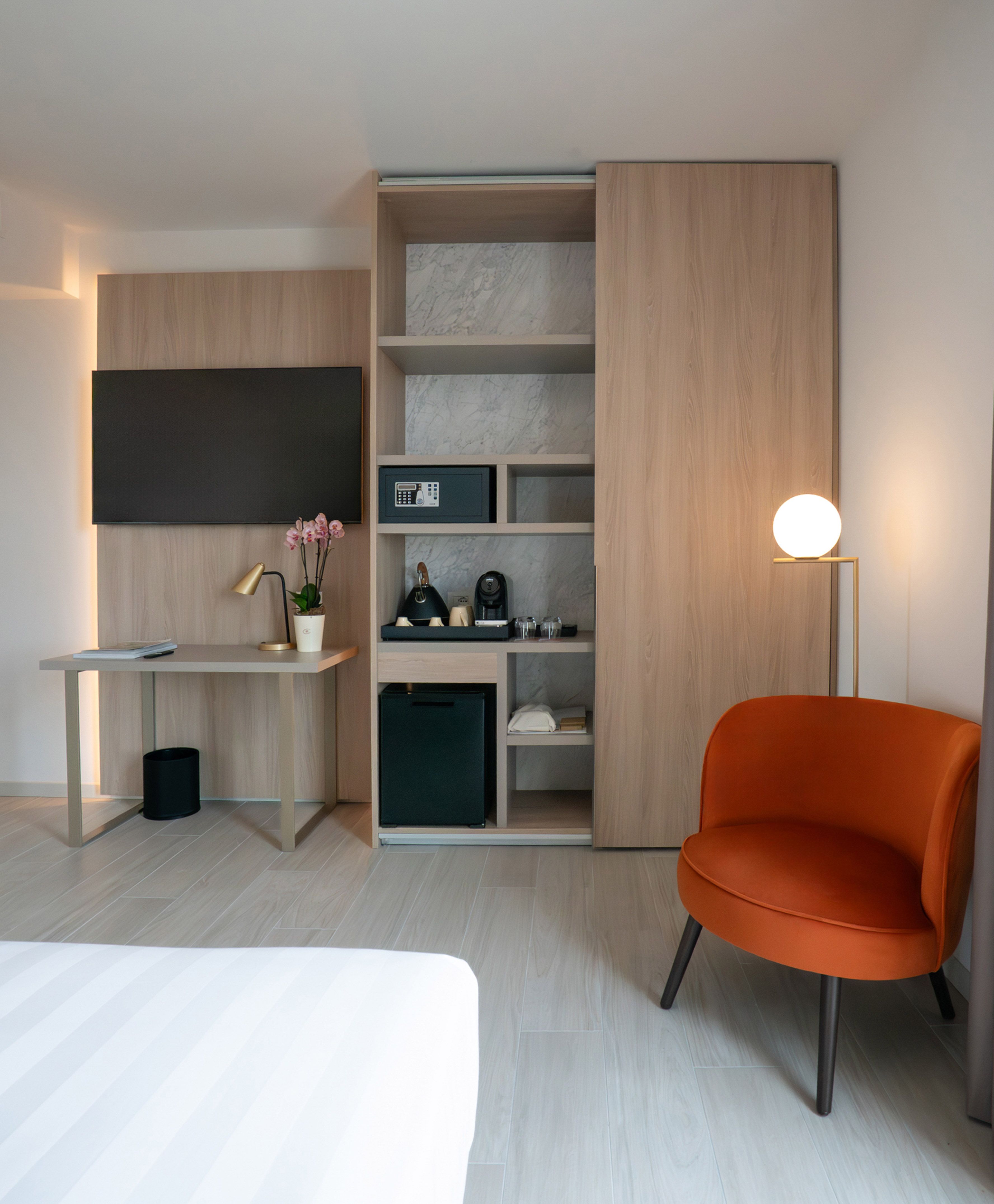 Modern hotel room: TV, desk, integrated wardrobe, minibar, orange armchair.