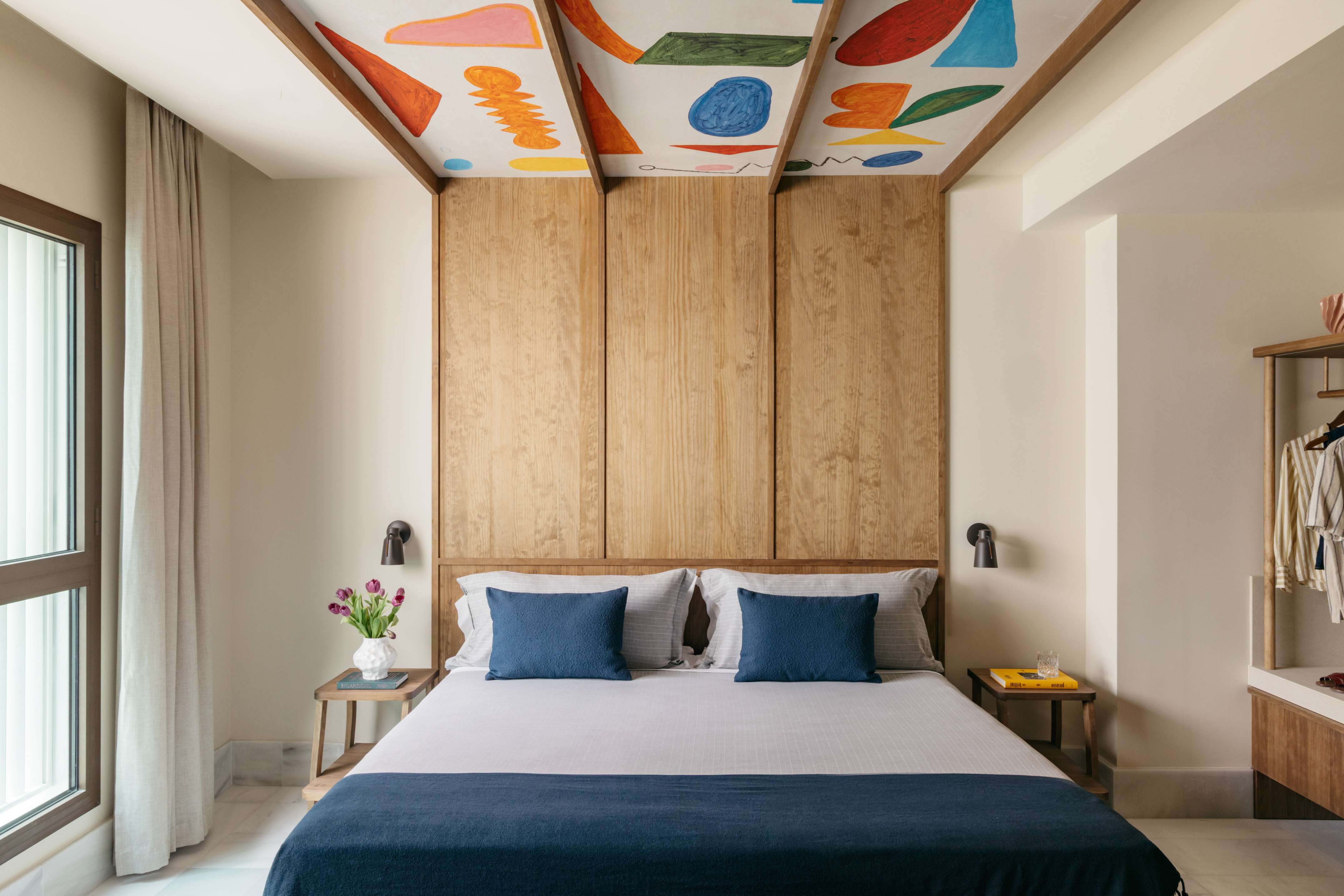 Modern bedroom with colorful abstract ceiling, wooden headboard, and blue bedding.