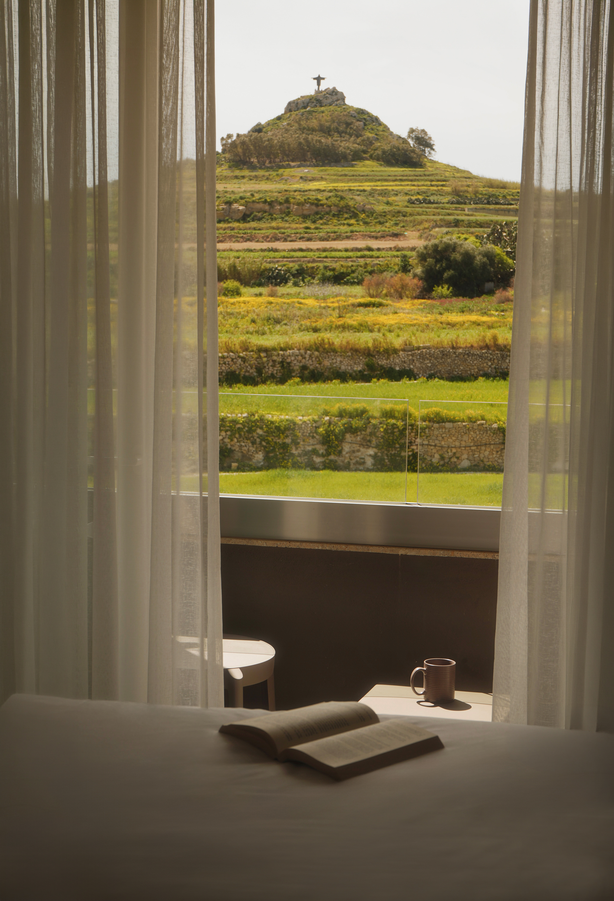 Bedroom view: terraced hills, hill-top statue, sheer curtains, open book, mug.