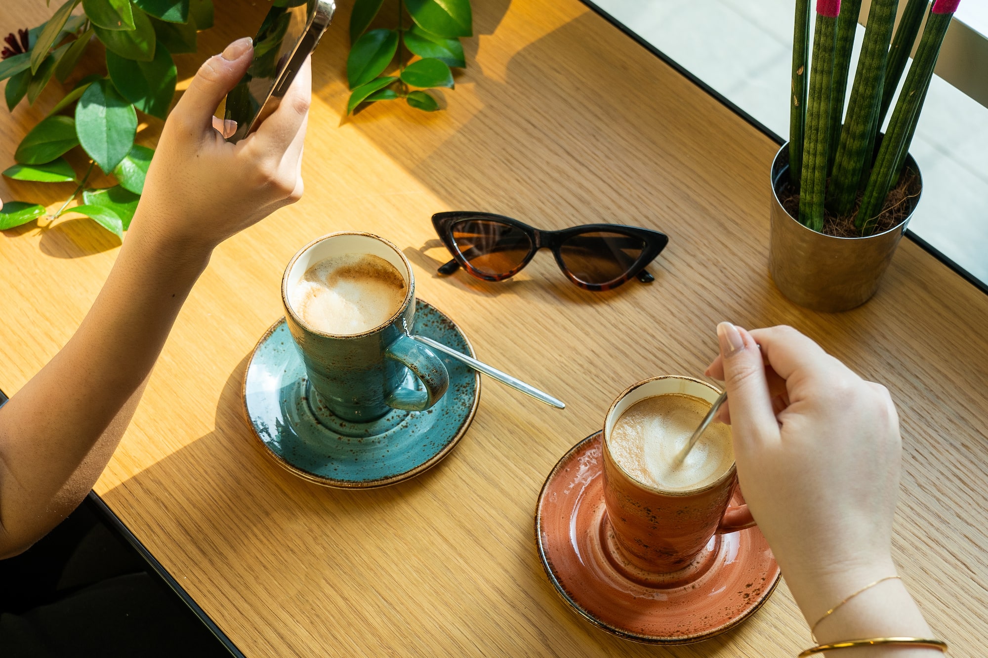 Dos cafés latte, manos con móvil, gafas de sol y plantas en mesa madera.