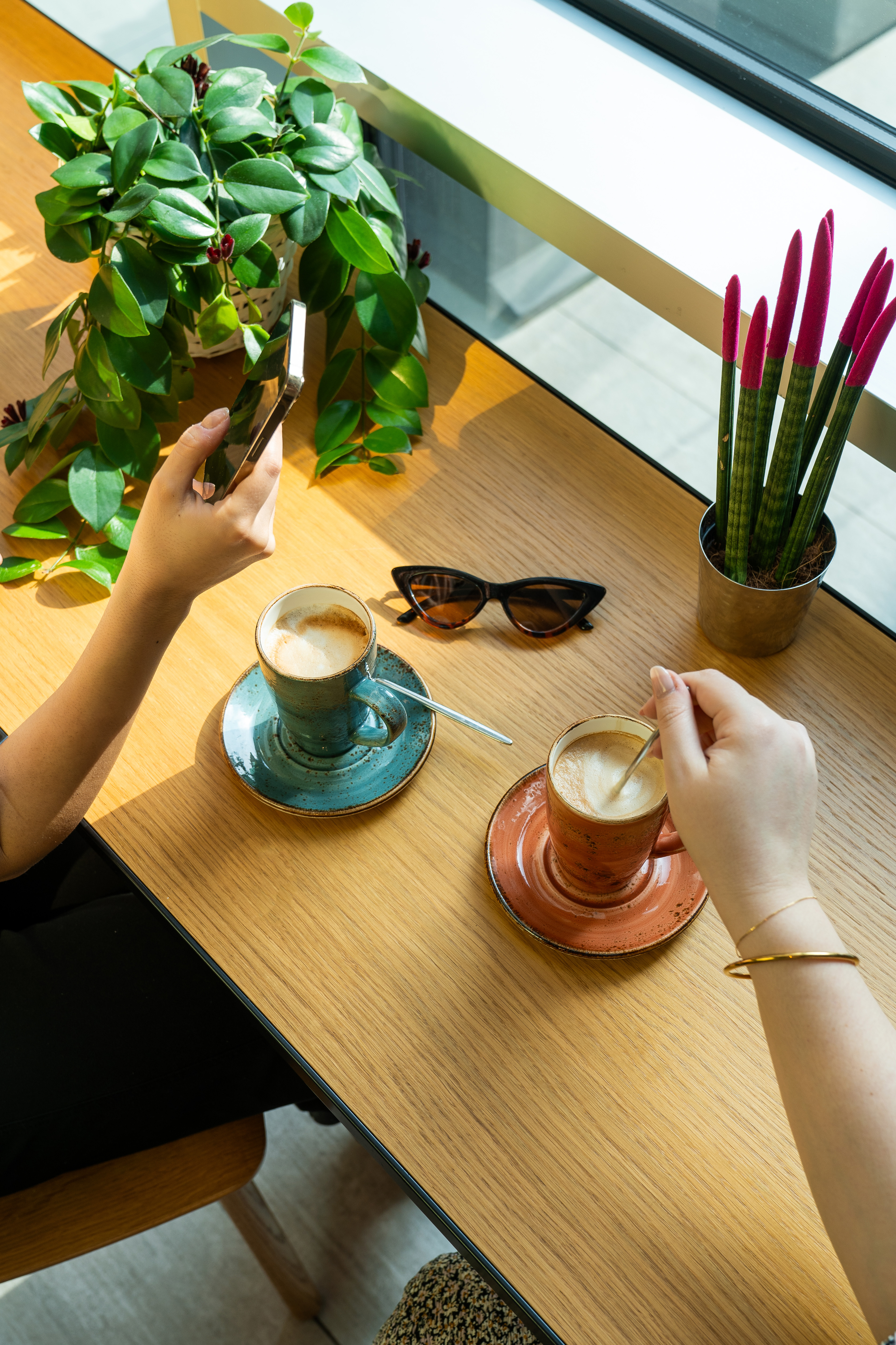 Dos cafés latte, manos con móvil, gafas de sol y plantas en mesa madera.