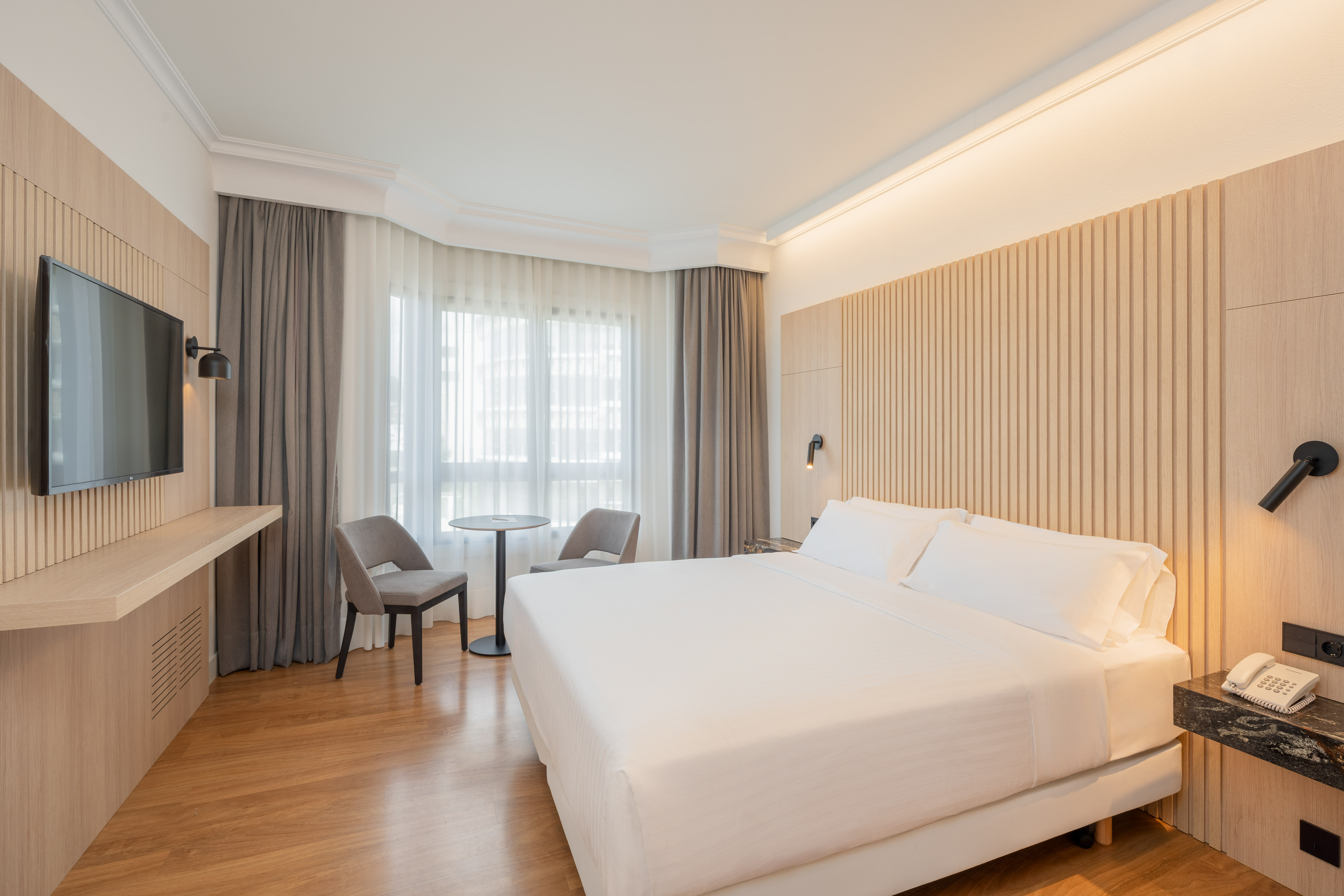 Modern hotel room with king bed, wood paneling, TV, and window seating.