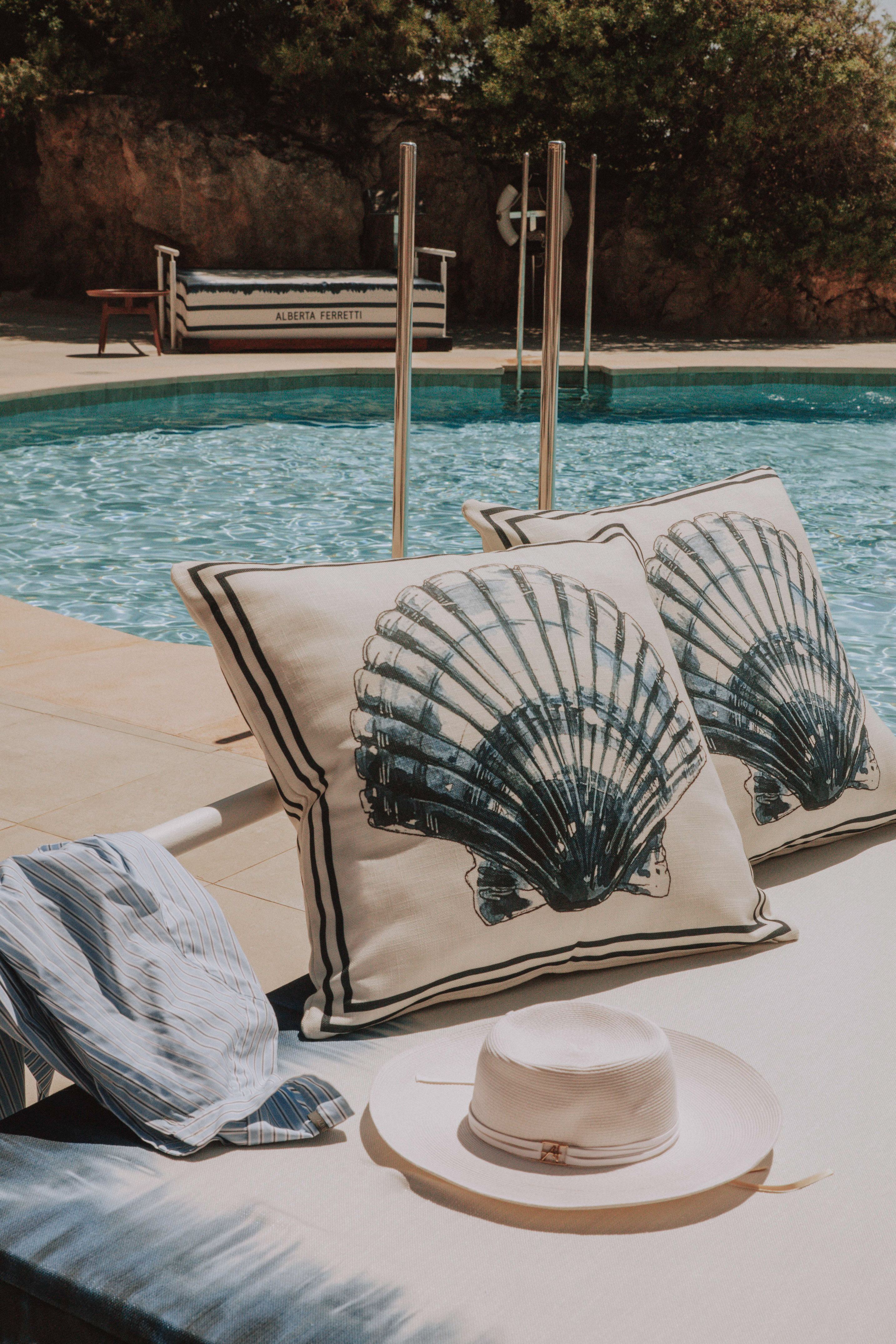 Summer poolside lounge with shell cushions, straw hat, and blue pool.