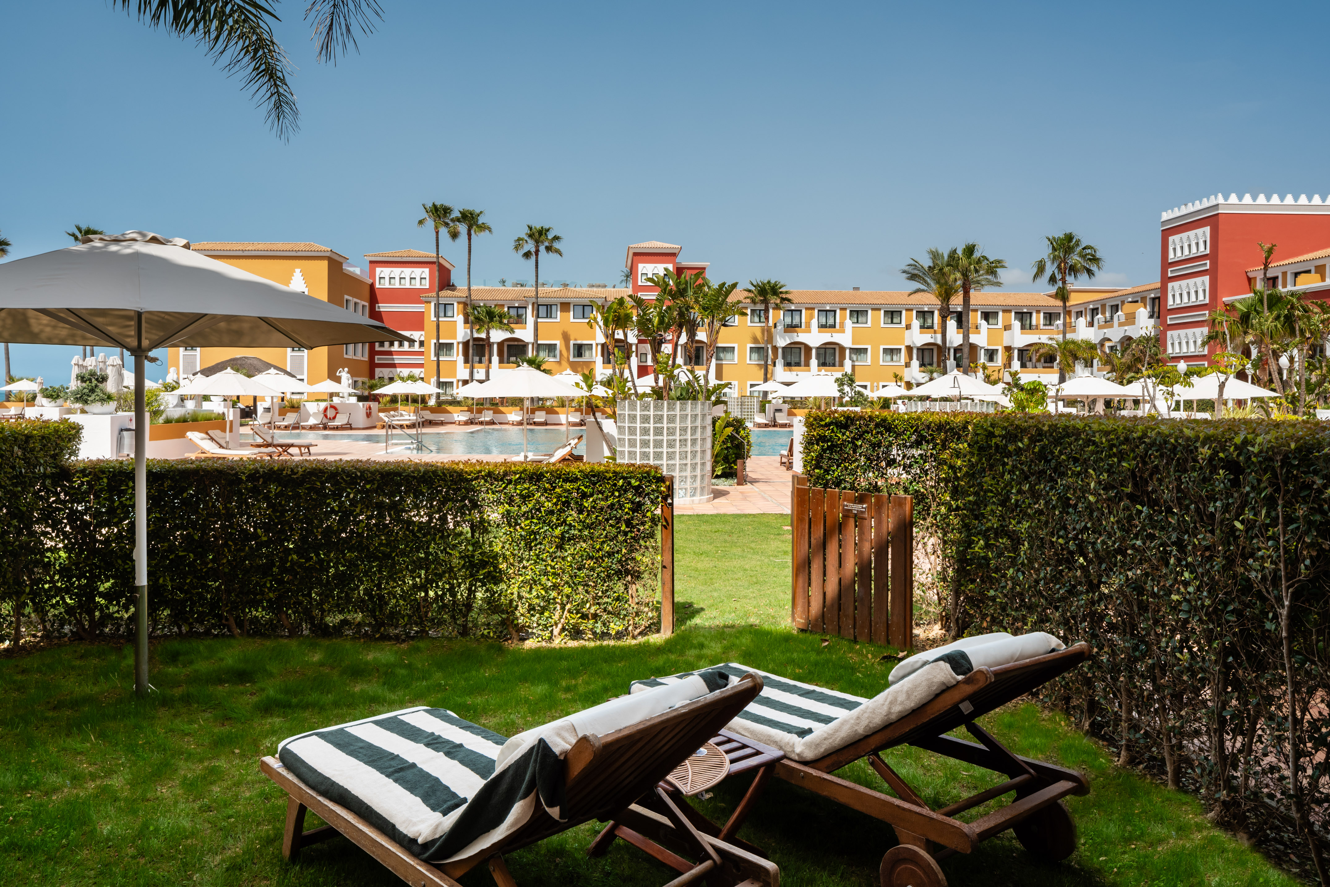 Sunny hotel resort pool with colorful building, palm trees, and lawn loungers.