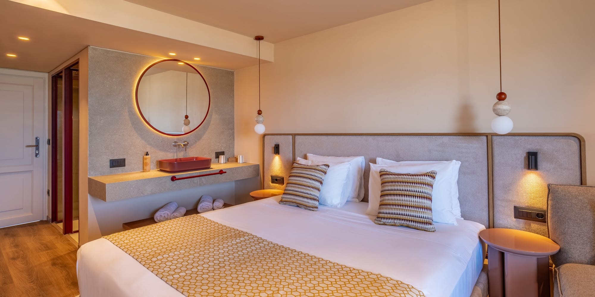 Luxurious modern hotel room with king bed, red vanity sink, and round backlit mirror.