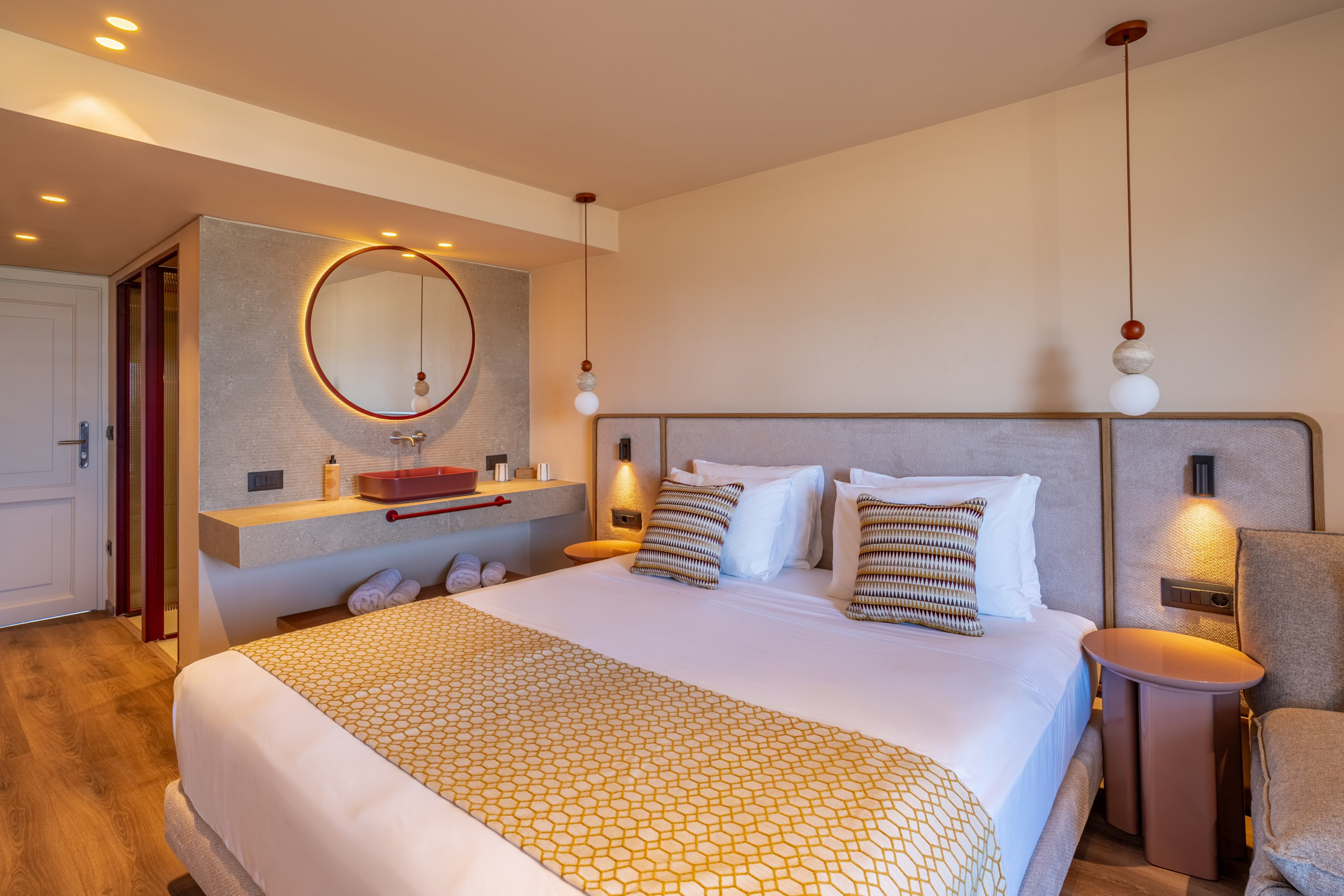 Luxurious modern hotel room with king bed, red vanity sink, and round backlit mirror.