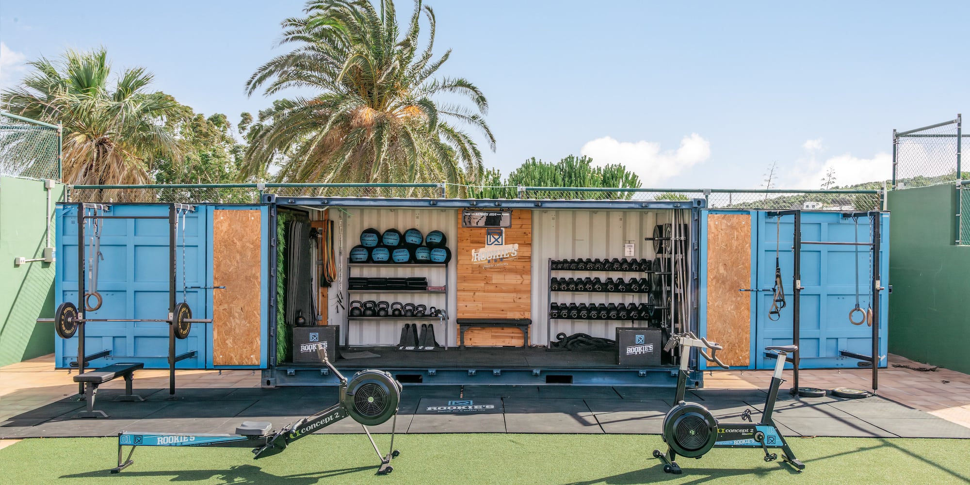 Blue shipping container outdoor gym with rowing machine, air bike, and weights.