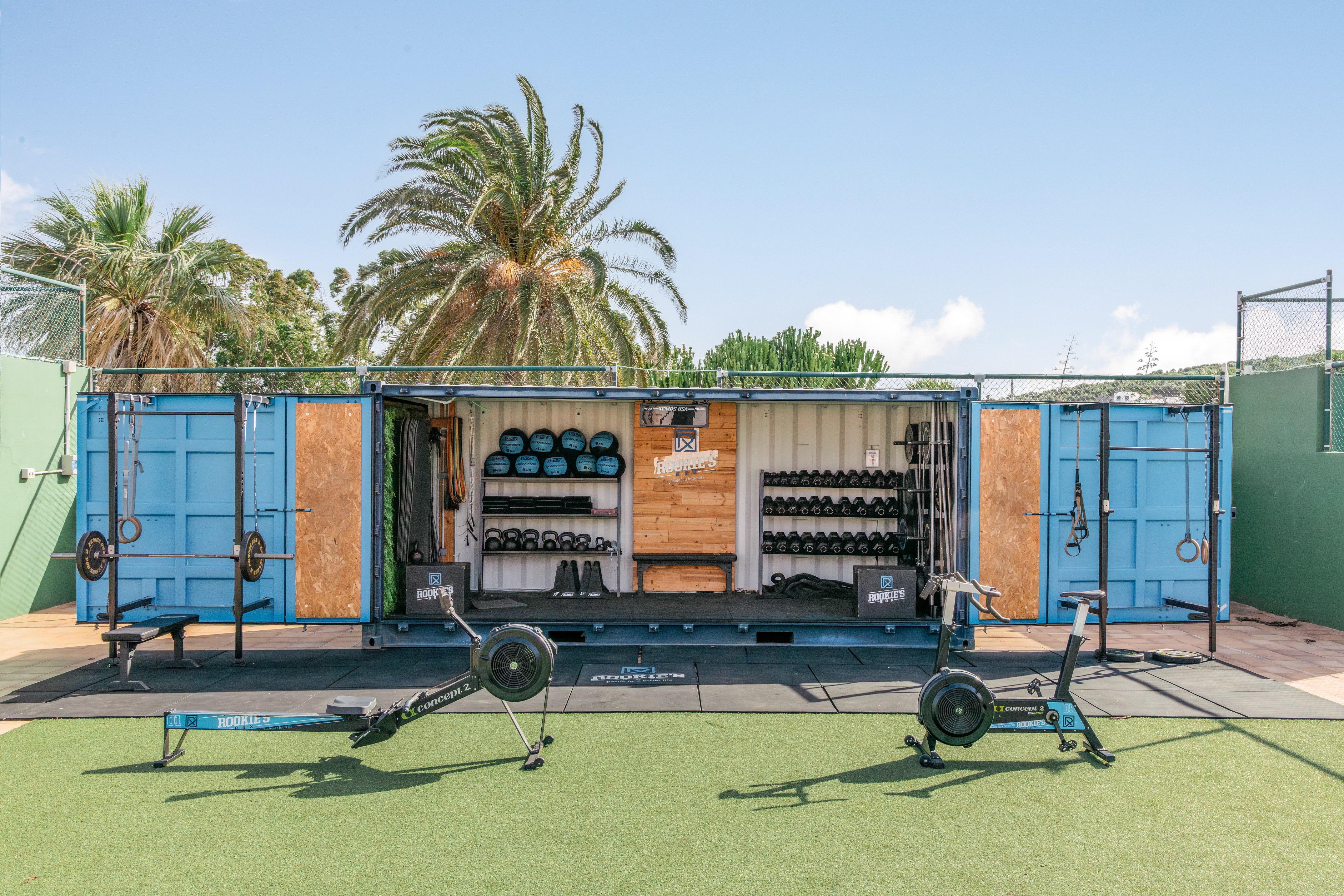 Blue shipping container outdoor gym with rowing machine, air bike, and weights.