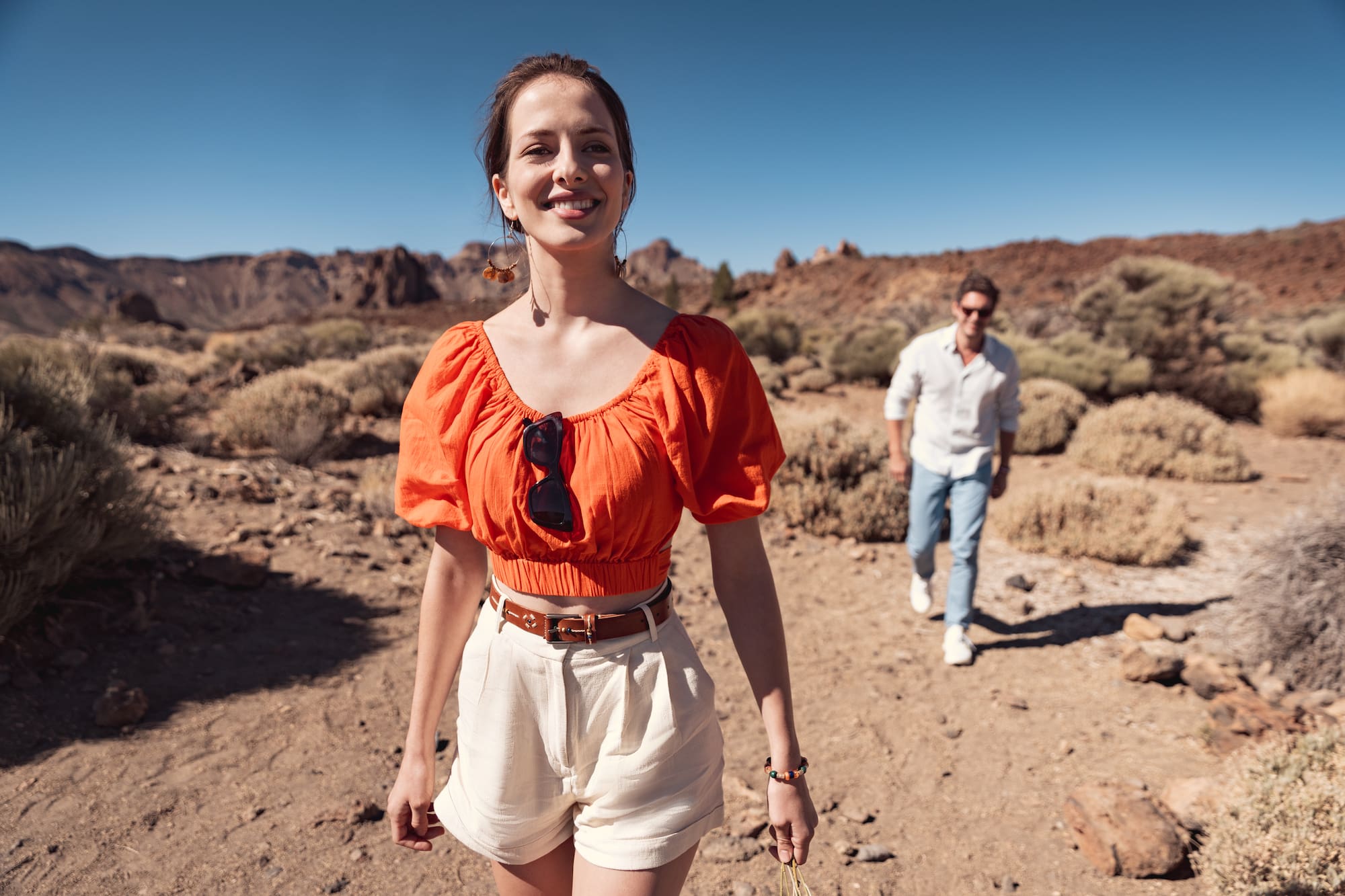 Smiling woman in orange top and white shorts hiking desert trail with man.