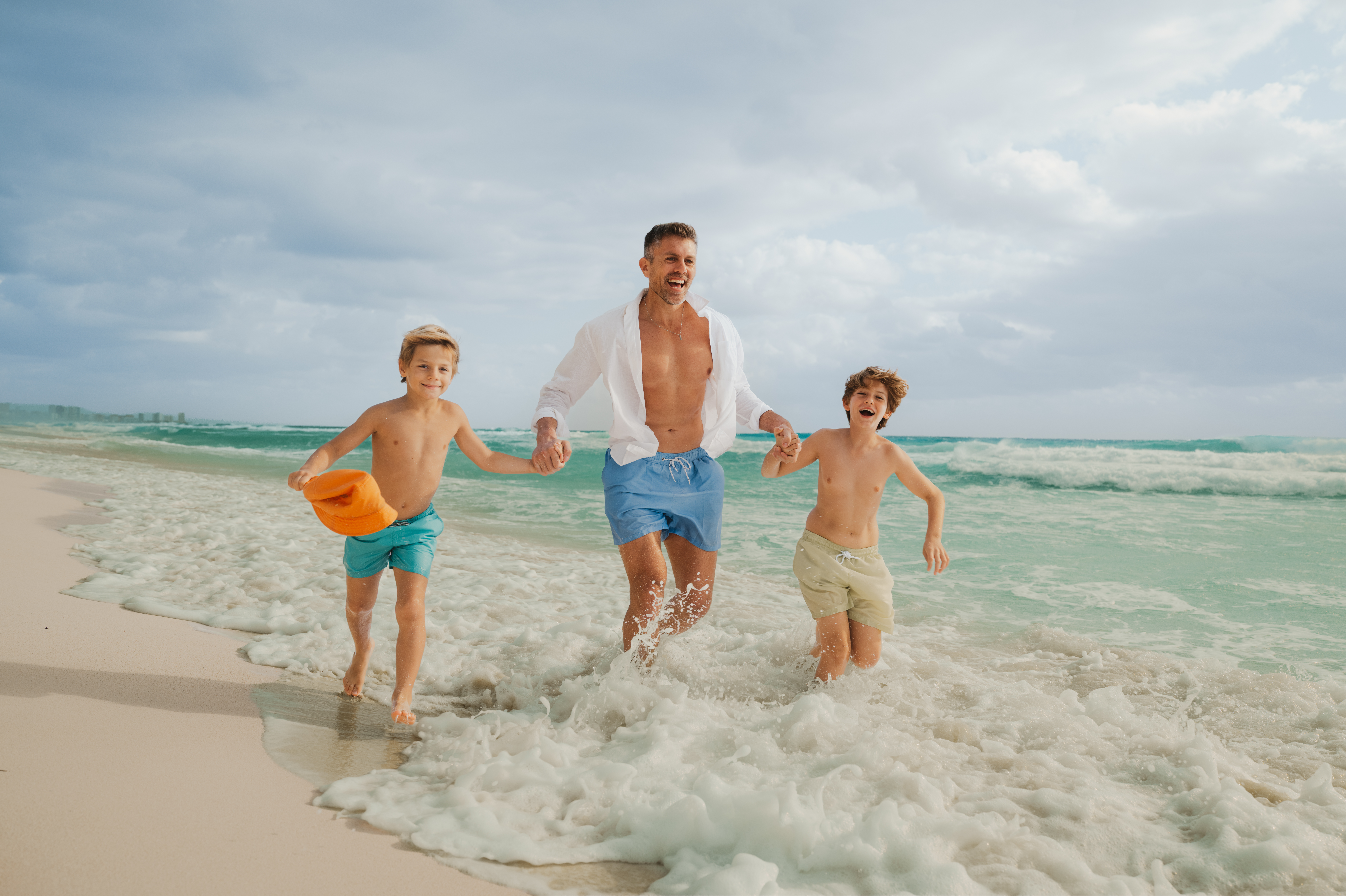 Padre y dos hijos corriendo de la mano en la playa, olas espumosas.