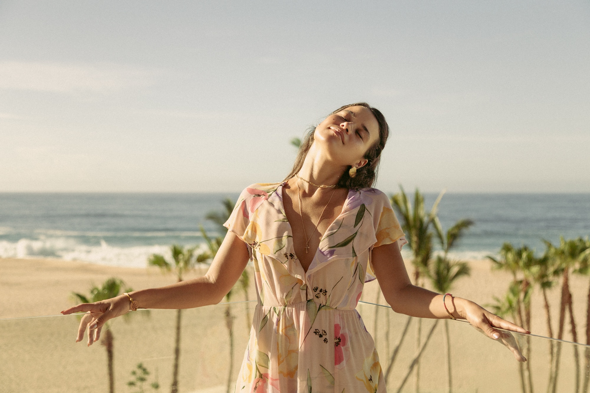 Mujer en vestido floral en balcón, disfrutando el sol, vista a playa tropical.