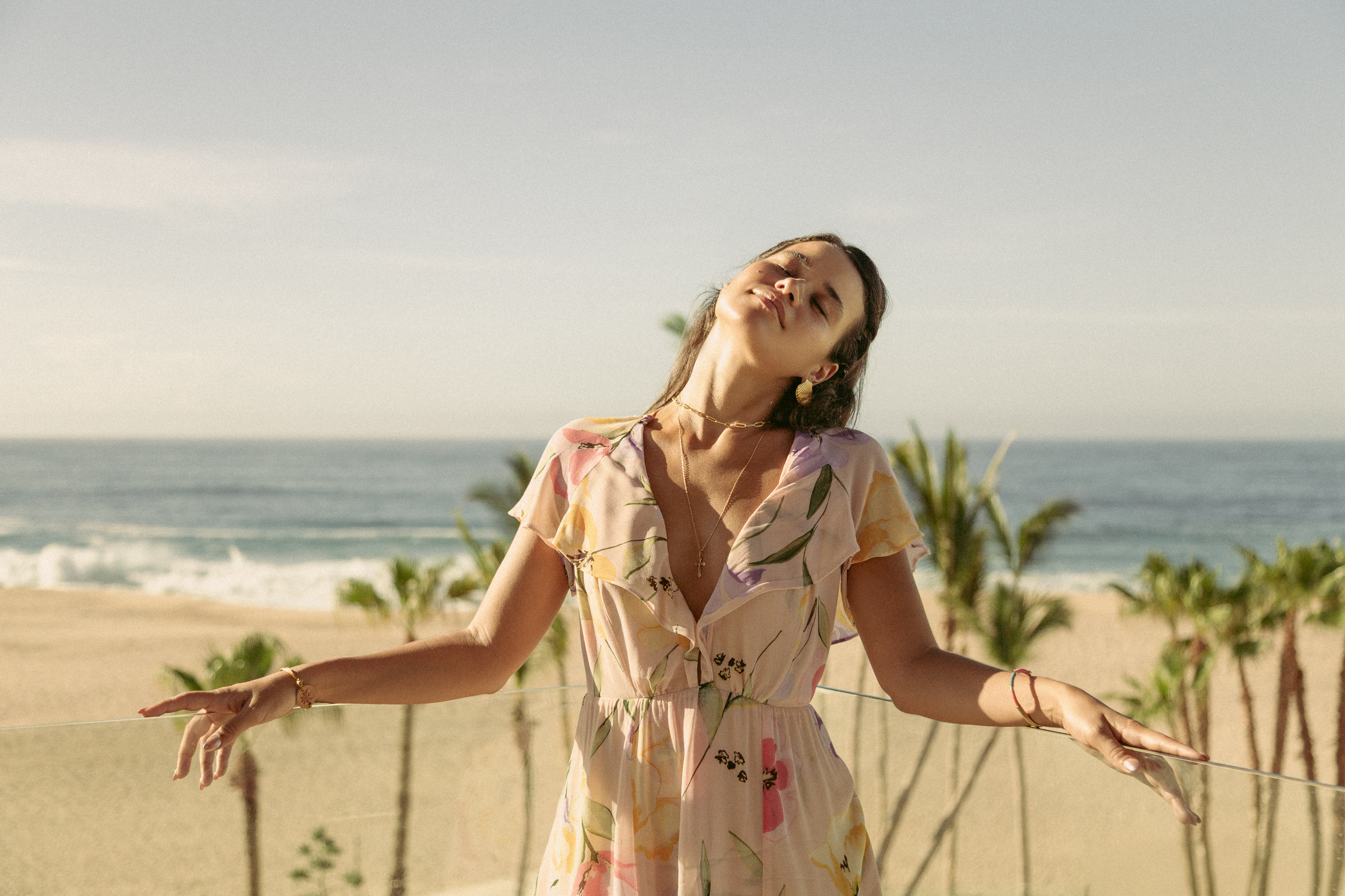 Mujer en vestido floral en balcón, disfrutando el sol, vista a playa tropical.