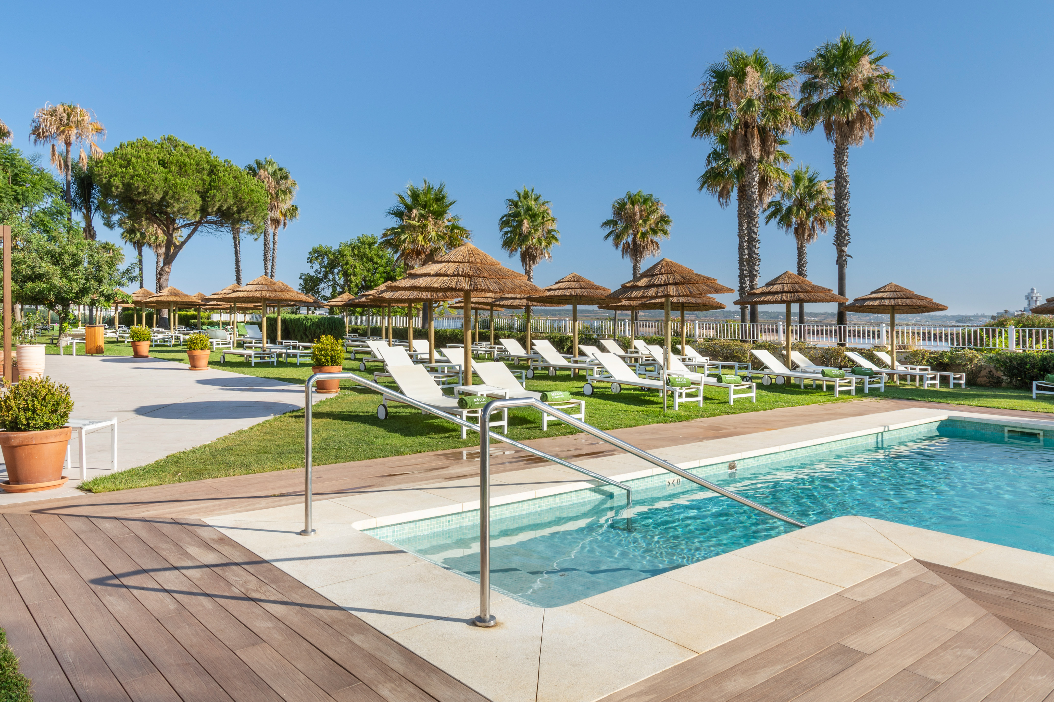 Resort swimming pool with sunbeds, thatched umbrellas, and palm trees.