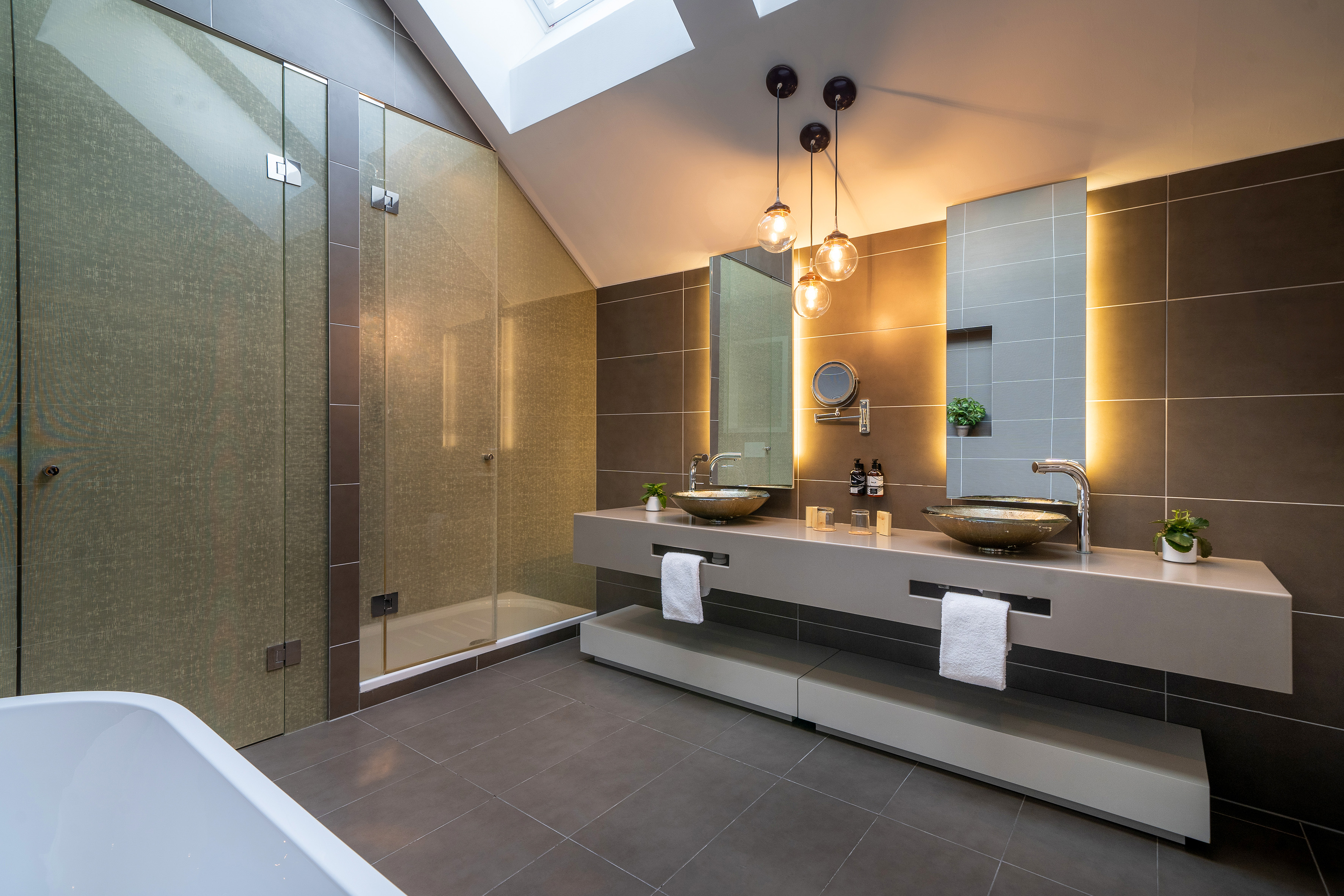 Modern bathroom with double vanity, glass shower, and skylight.