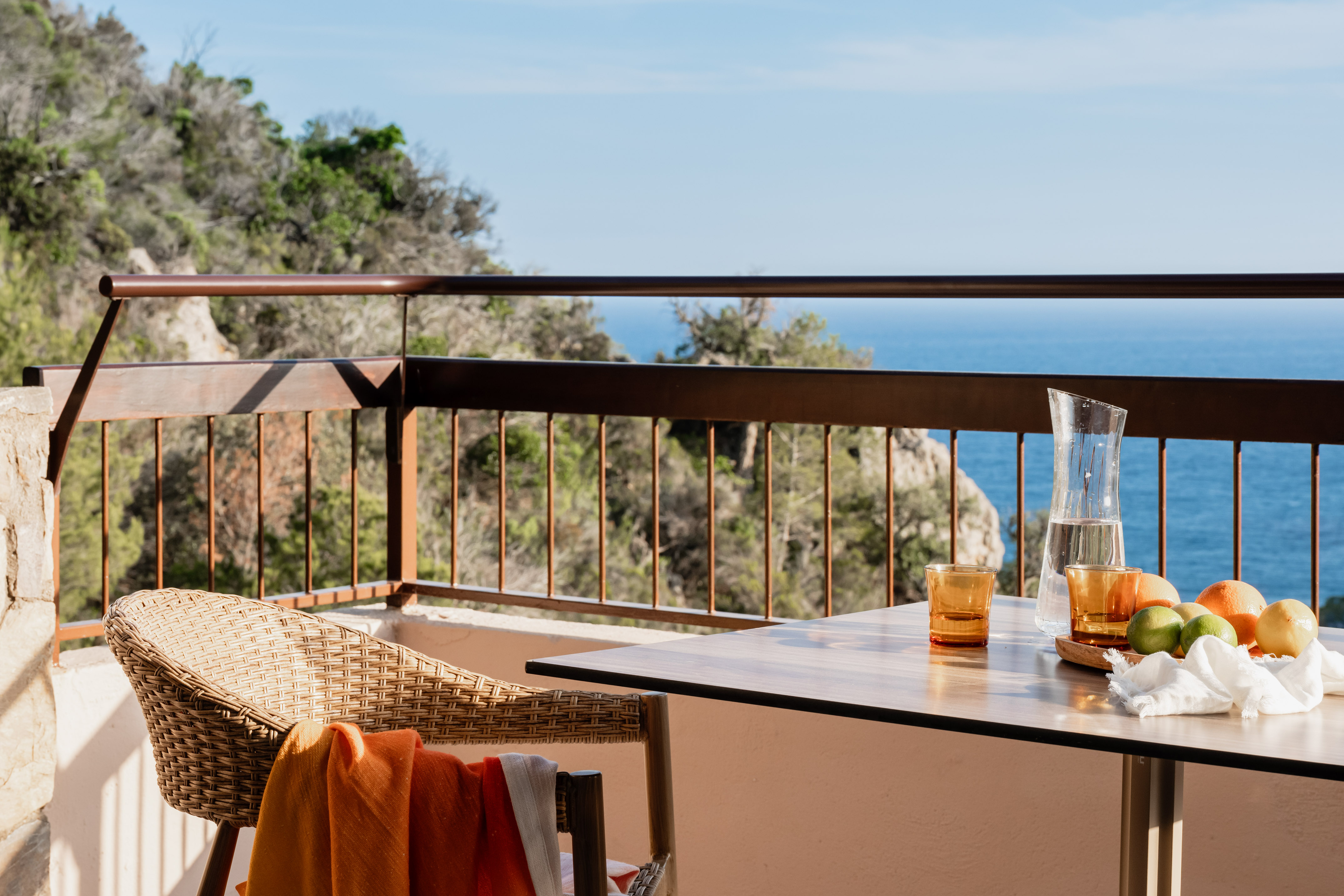 Sunny balcony overlooking the sea, table with water, fruit, and wicker chair.