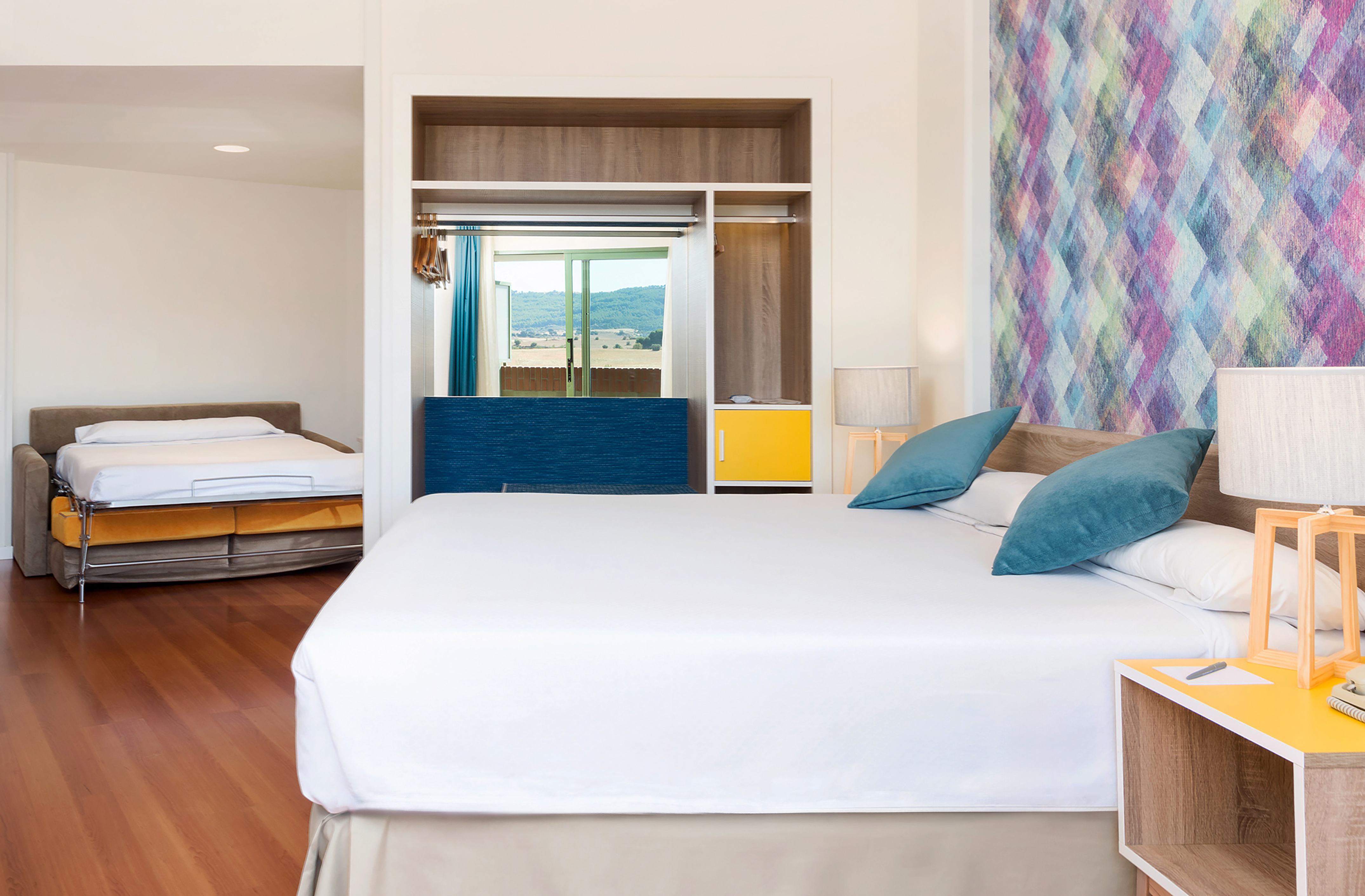 Modern hotel suite with large bed, pull-out sofa, colorful wall, scenic view.