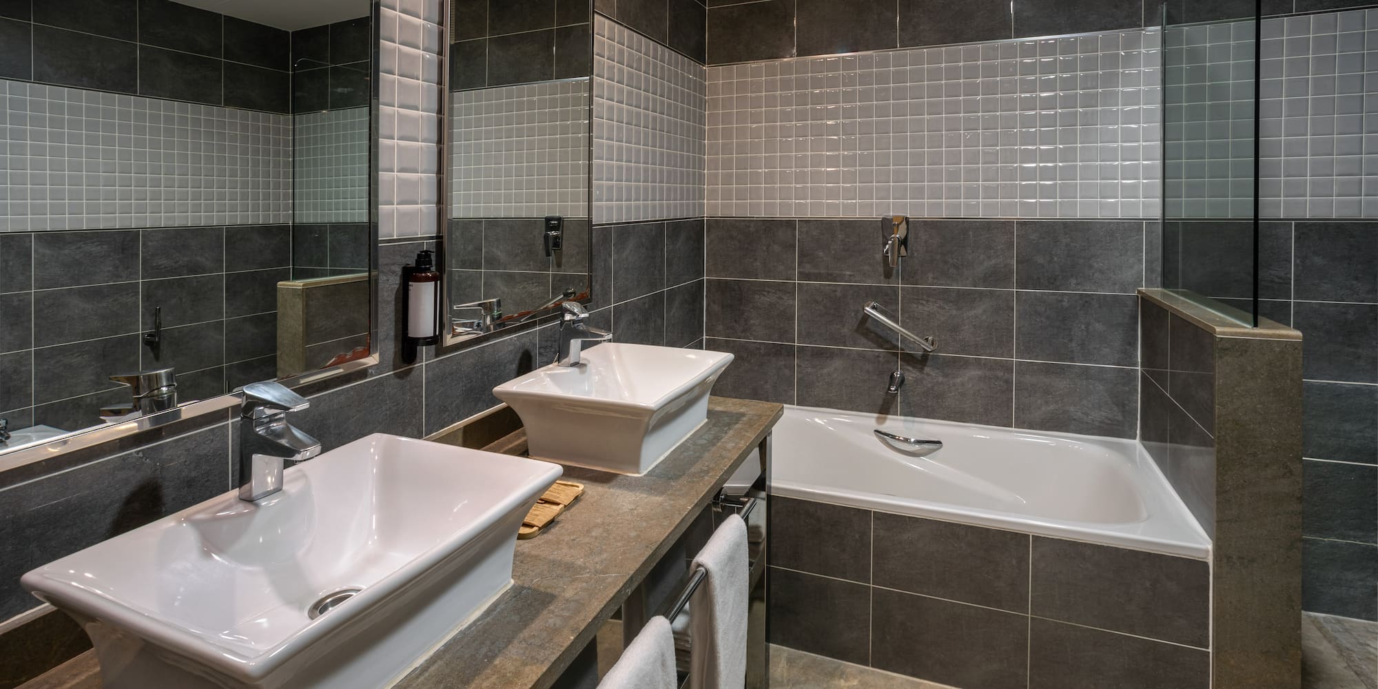 Modern bathroom with dual vessel sinks, bathtub, grey tiles, and large mirror.