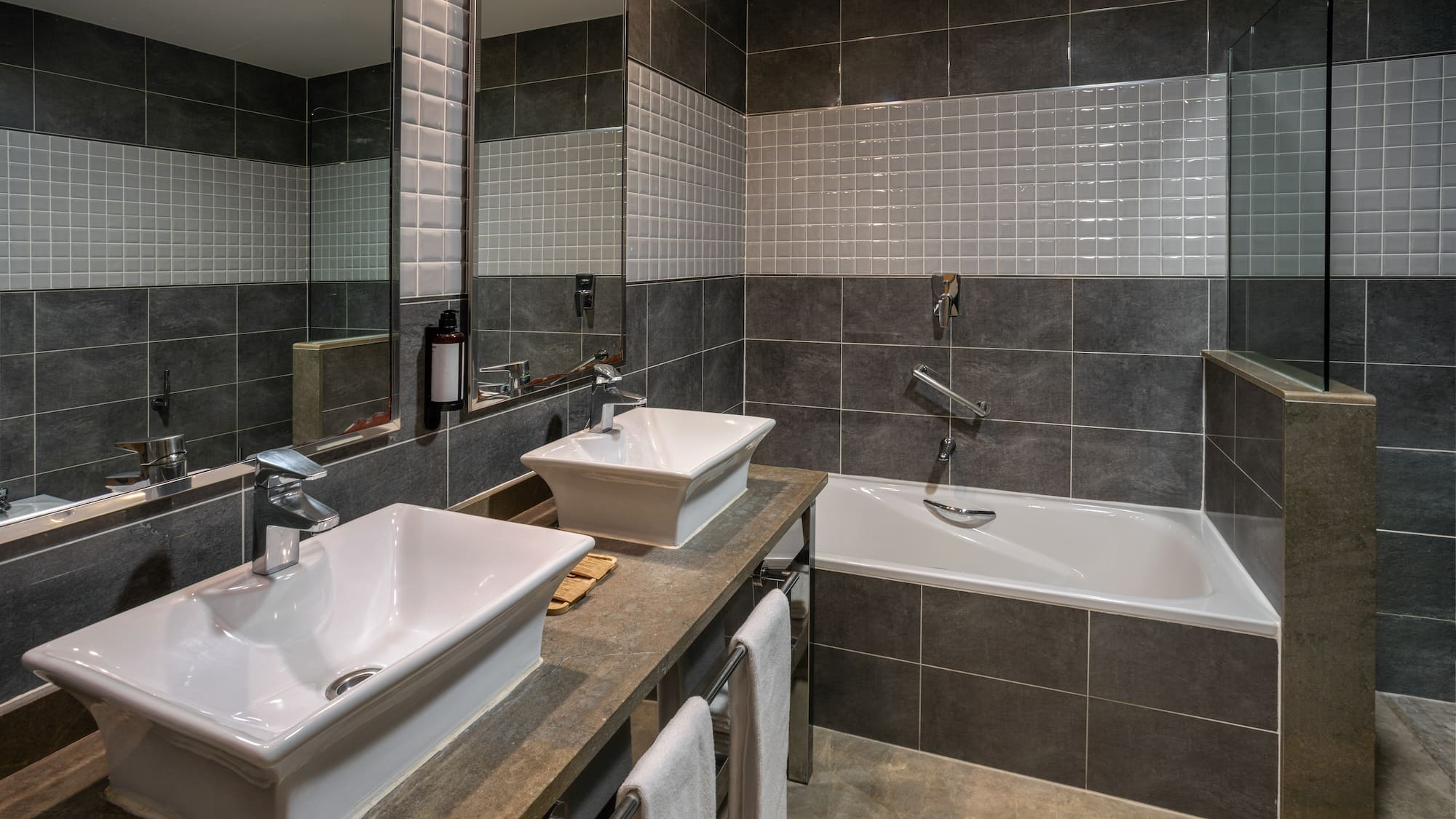 Modern bathroom with dual vessel sinks, bathtub, grey tiles, and large mirror.