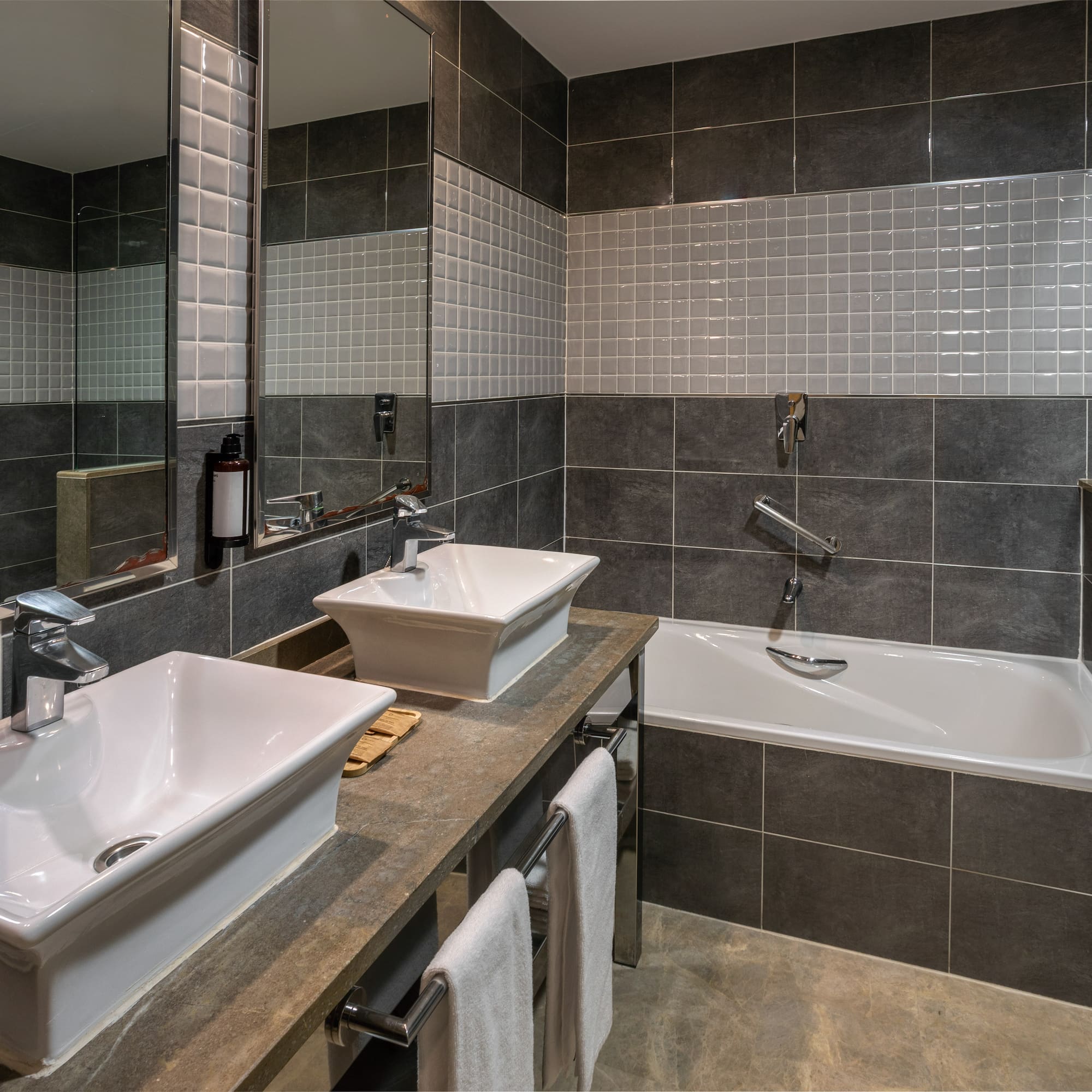 Modern bathroom with dual vessel sinks, bathtub, grey tiles, and large mirror.