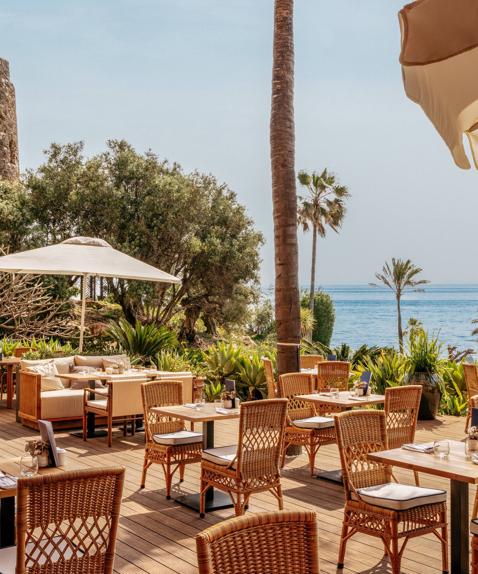 Outdoor restaurant terrace with sea view, palm trees, and historic stone tower.