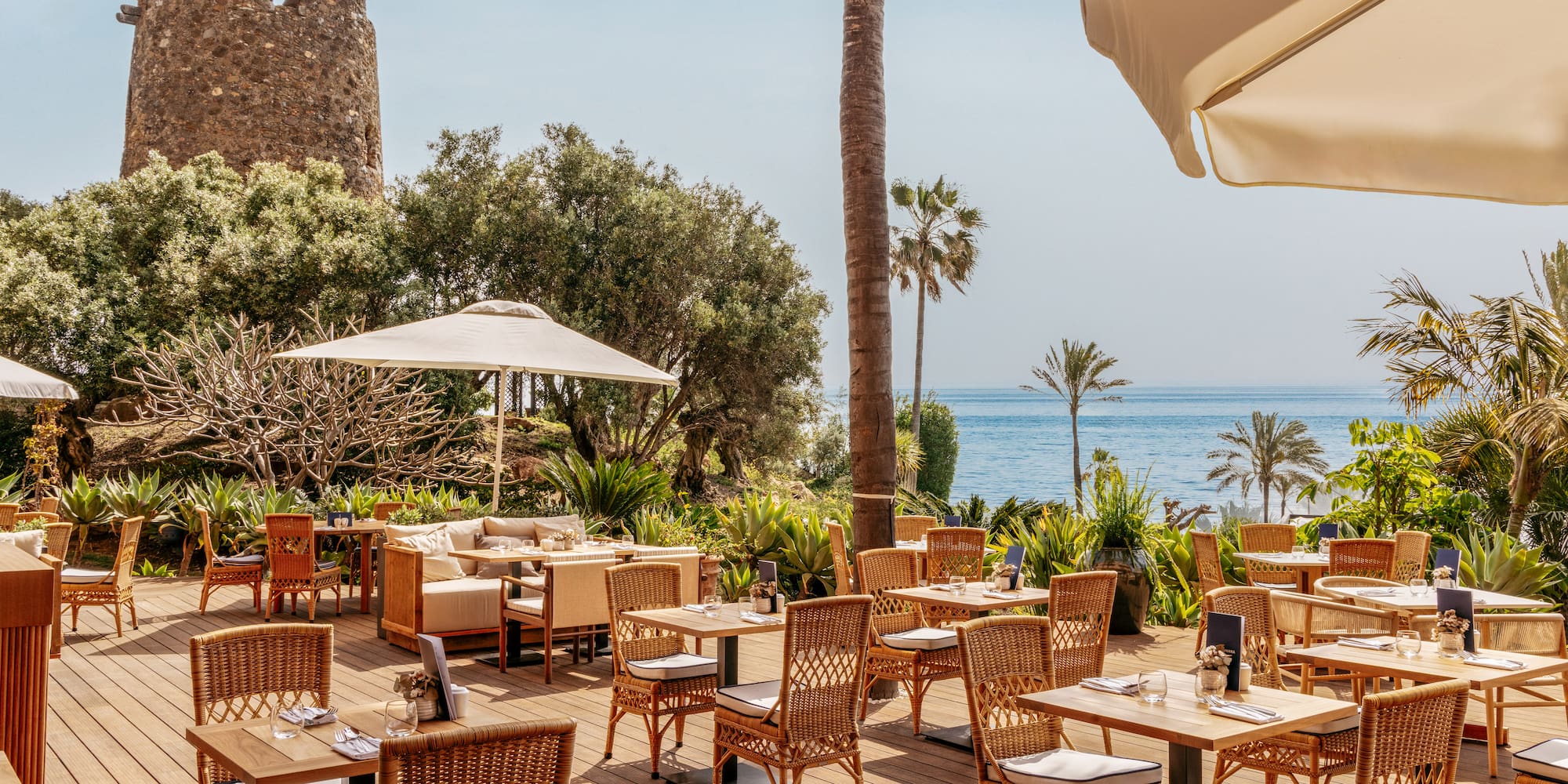 Coastal restaurant patio with sea view, historic stone tower, and palm trees.