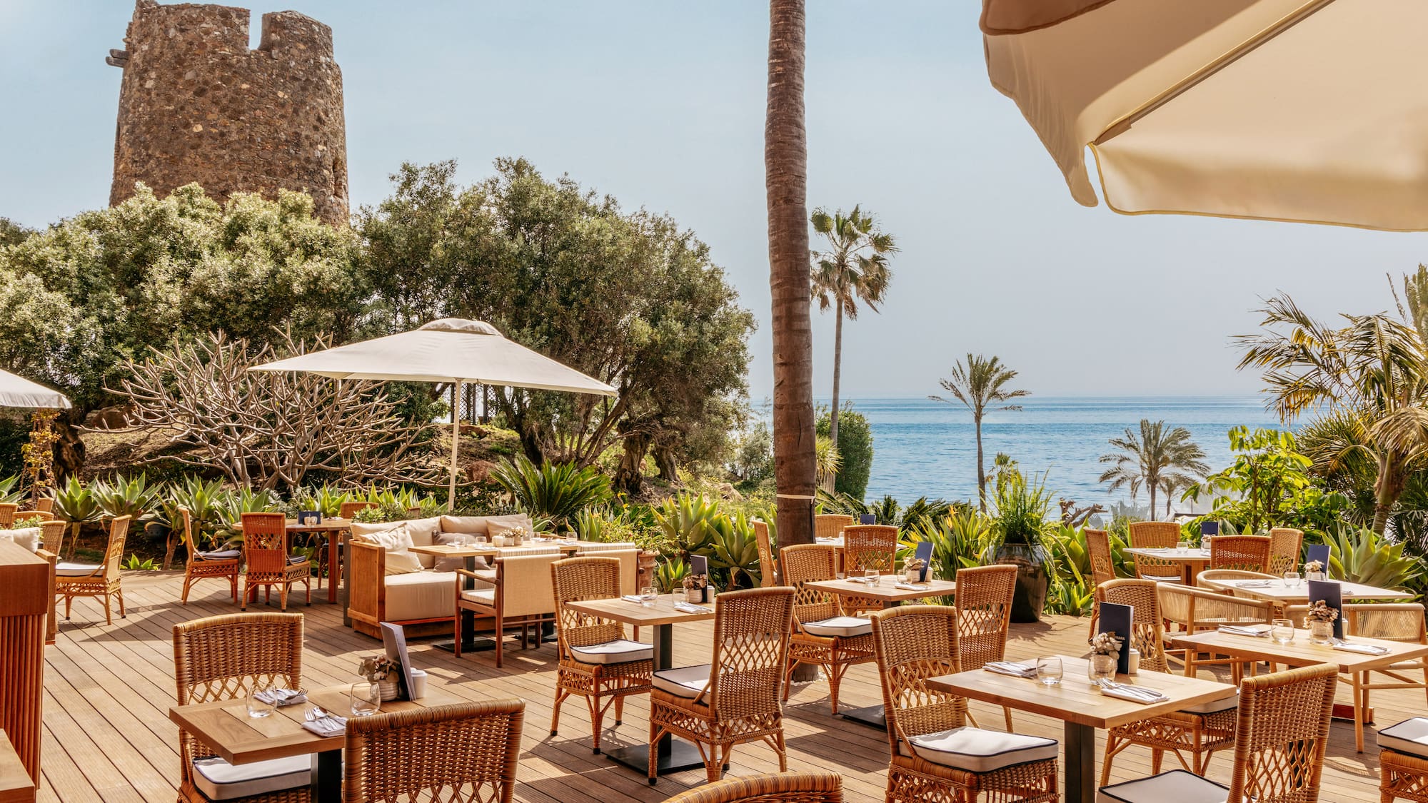 Coastal restaurant patio with sea view, historic stone tower, and palm trees.