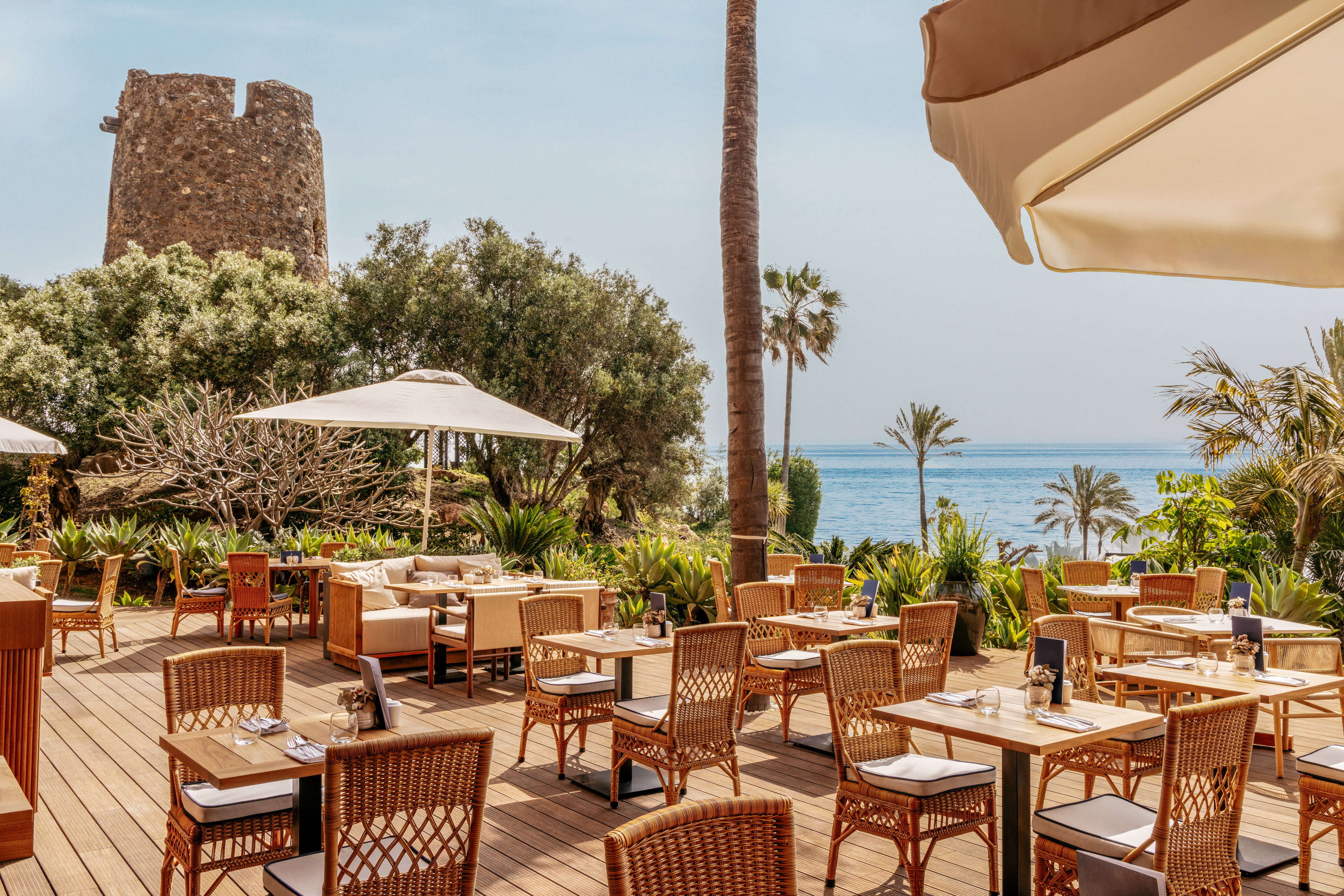 Coastal restaurant patio with sea view, historic stone tower, and palm trees.