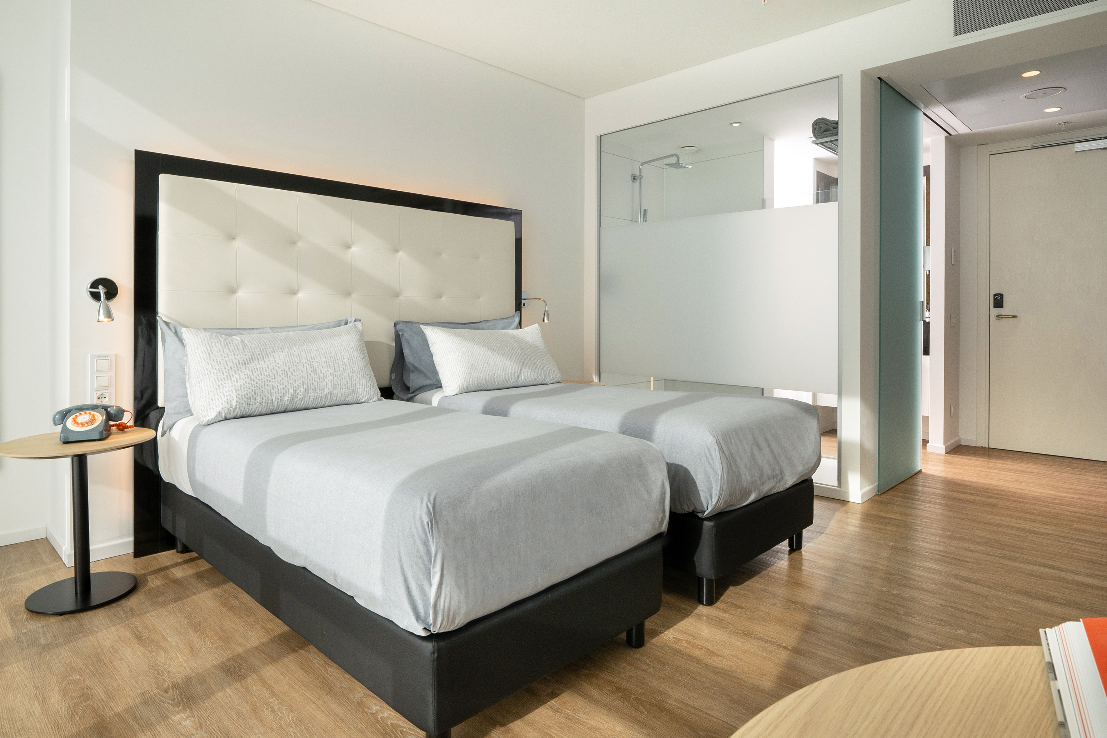 Modern hotel room with two twin beds, wood floor, and glass shower.