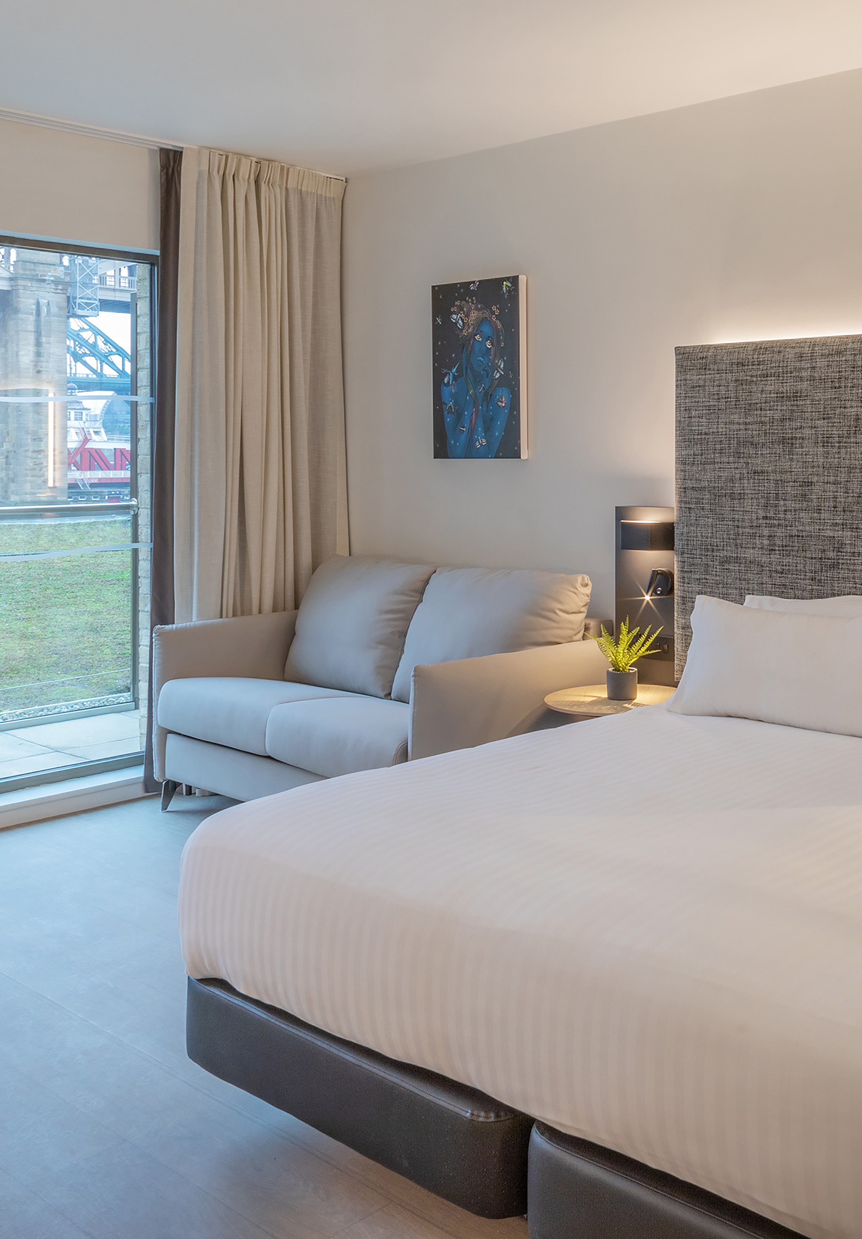 Modern hotel bedroom with white bed, sofa, abstract art, and bridge view.