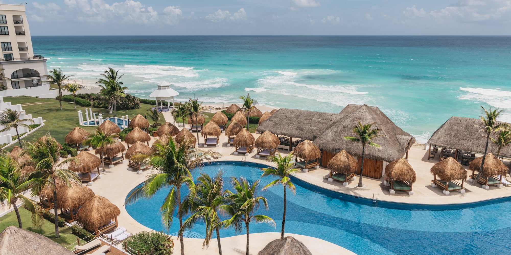 Oceanfront resort pool with palm trees, thatched cabanas, and turquoise ocean.