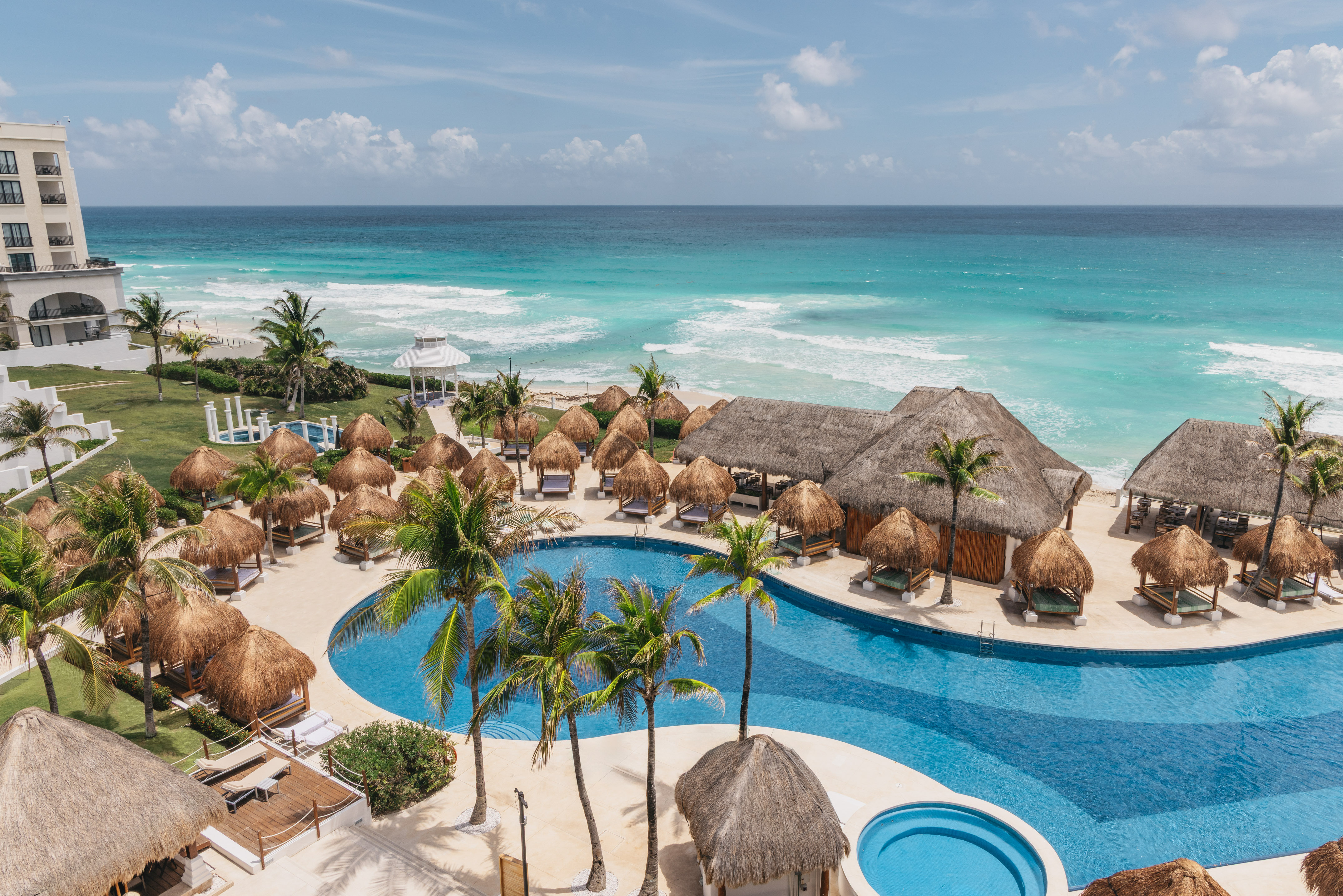 Oceanfront resort pool with palm trees, thatched cabanas, and turquoise ocean.