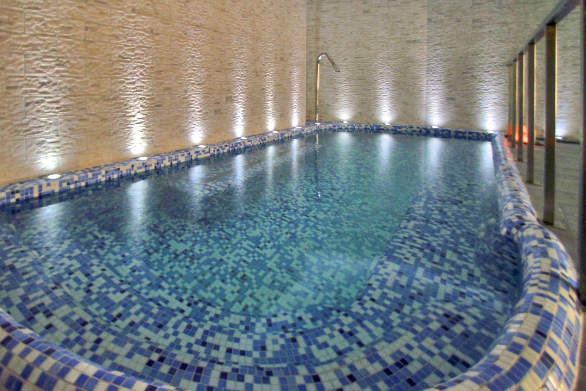 Indoor spa pool with blue mosaic tiles, textured wall, and accent lighting.