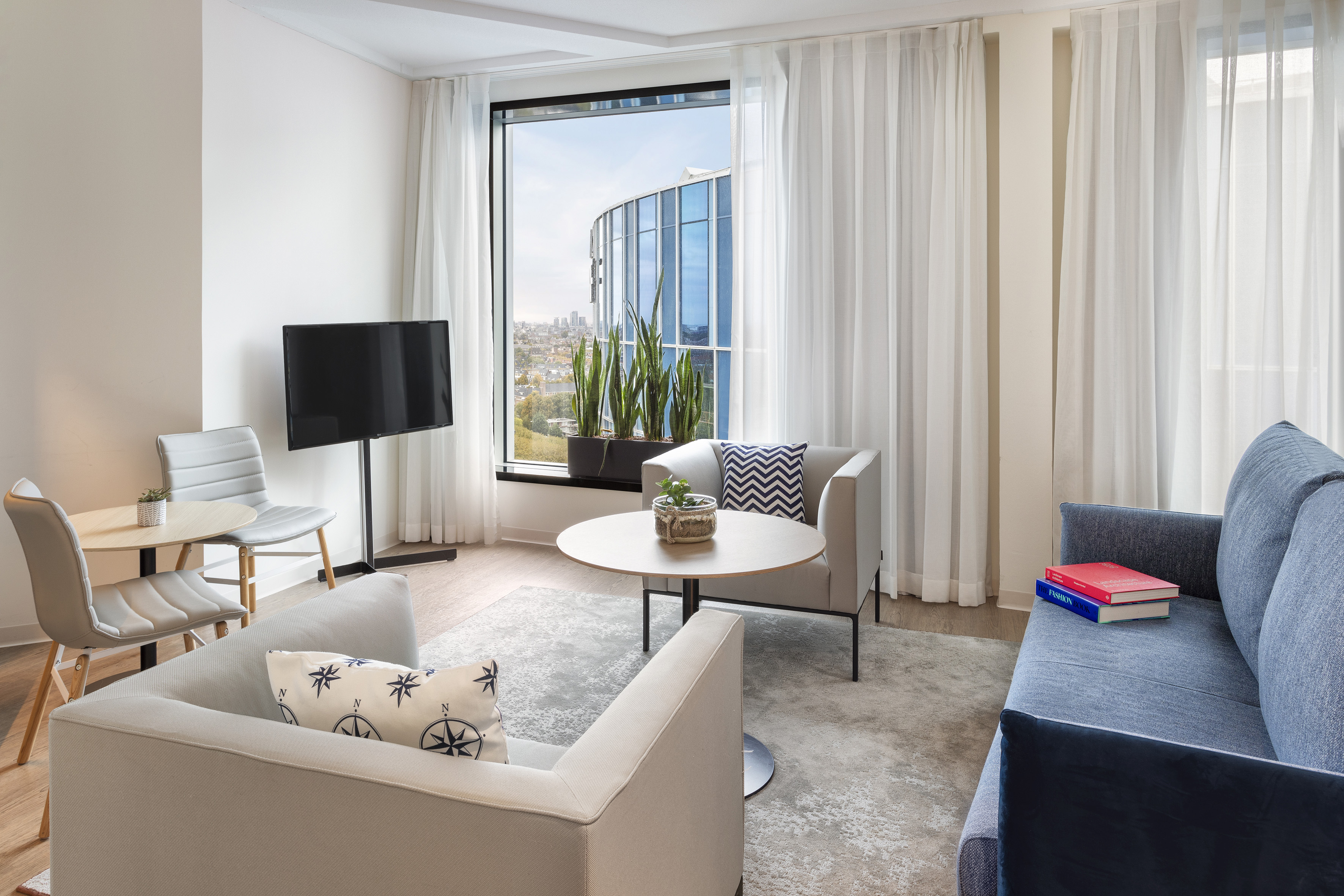 Contemporary hotel suite living room with city view, sofa, armchairs, and TV.