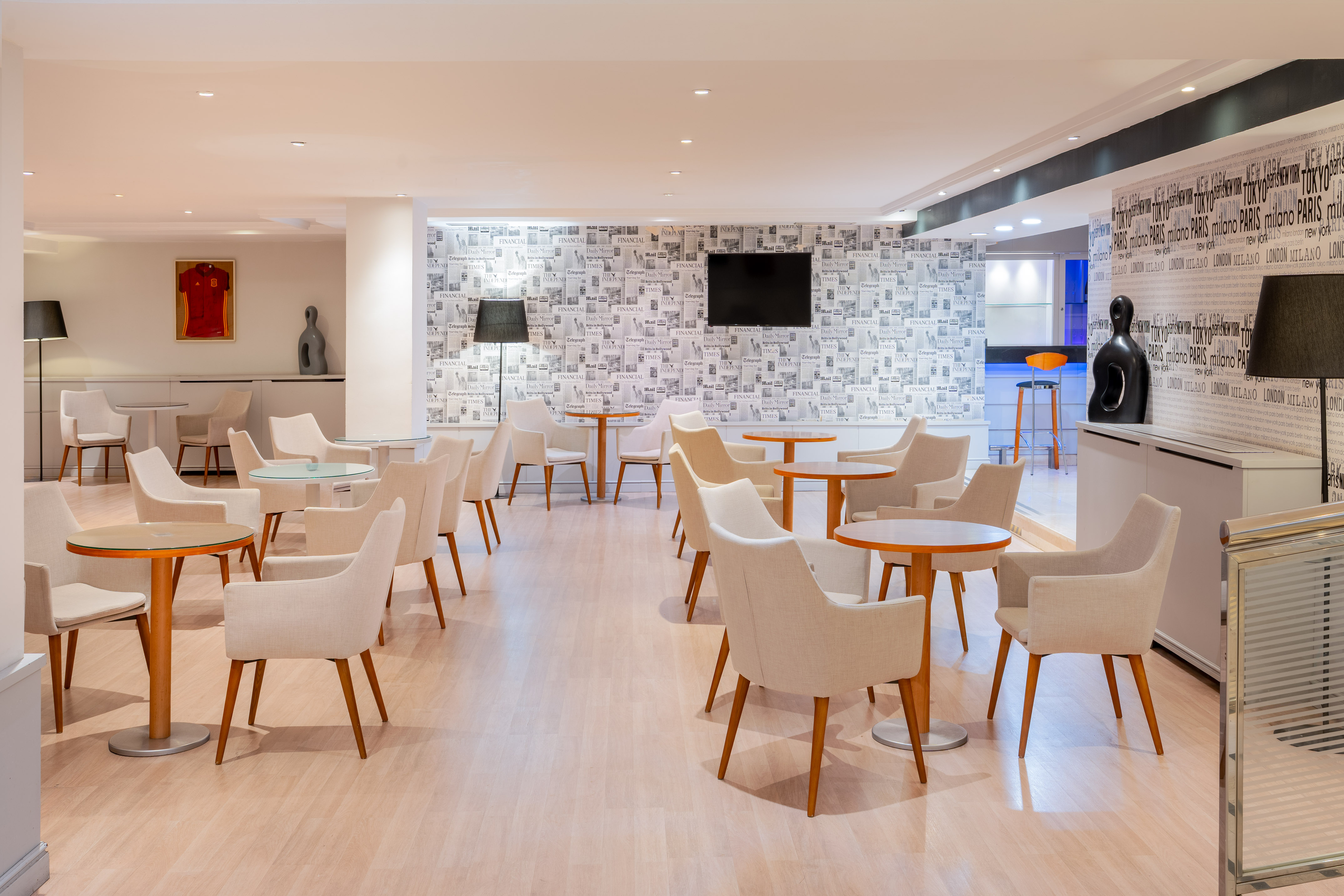 Hotel lounge with cream chairs, round tables, and newspaper-themed wallpaper.