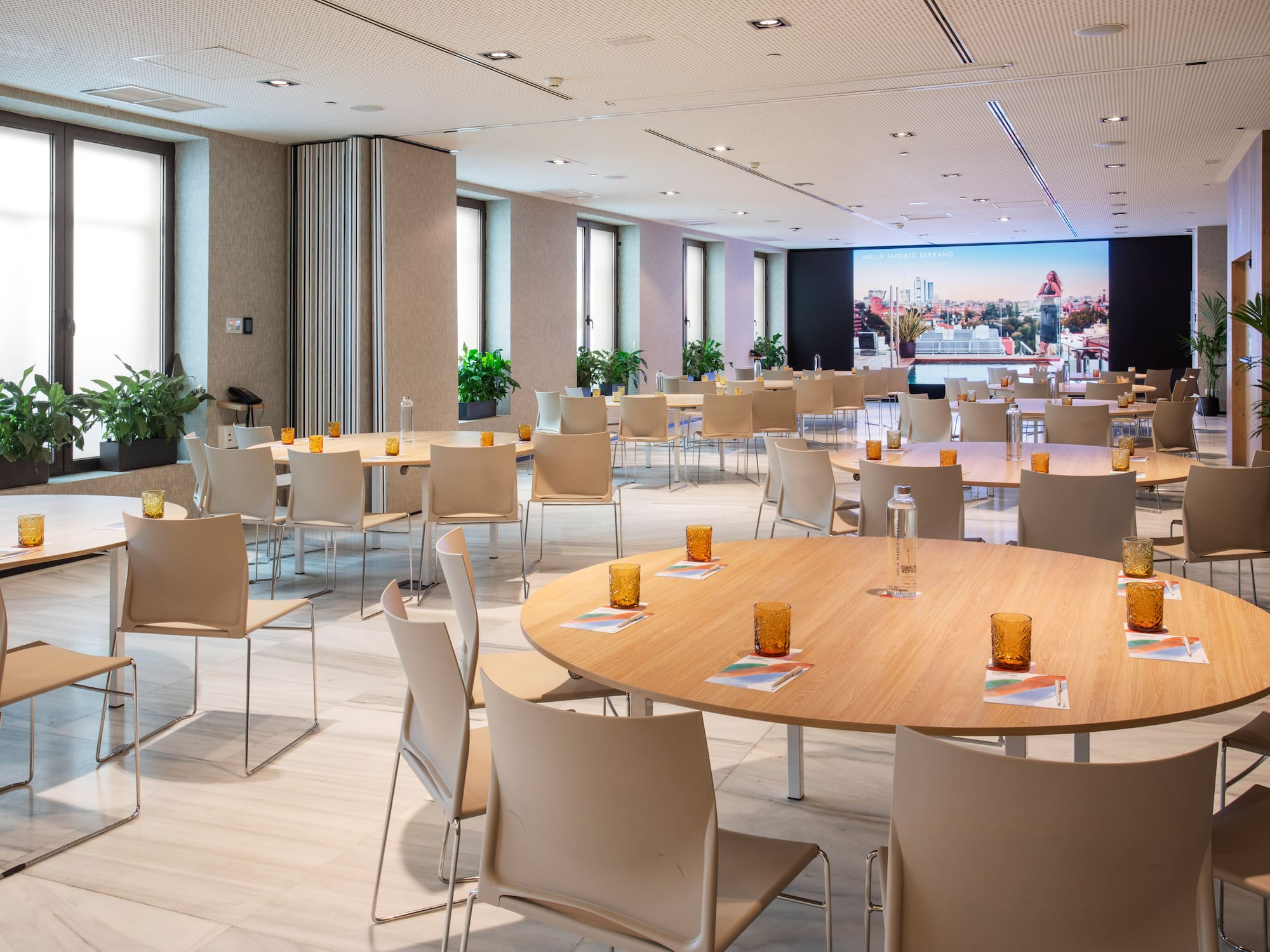 Modern hotel meeting space with round tables, chairs, windows, and large screen.