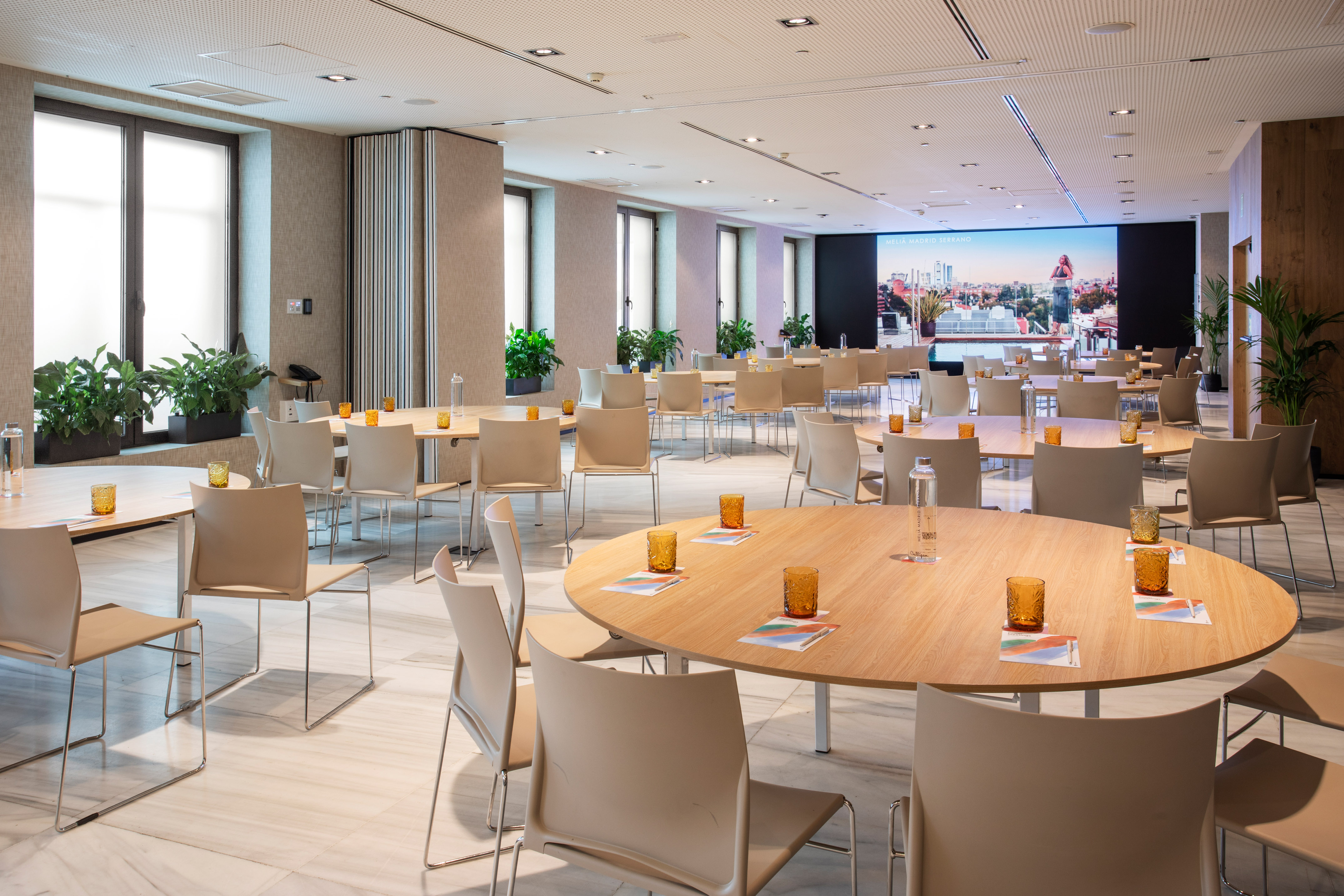 Modern hotel meeting space with round tables, chairs, windows, and large screen.