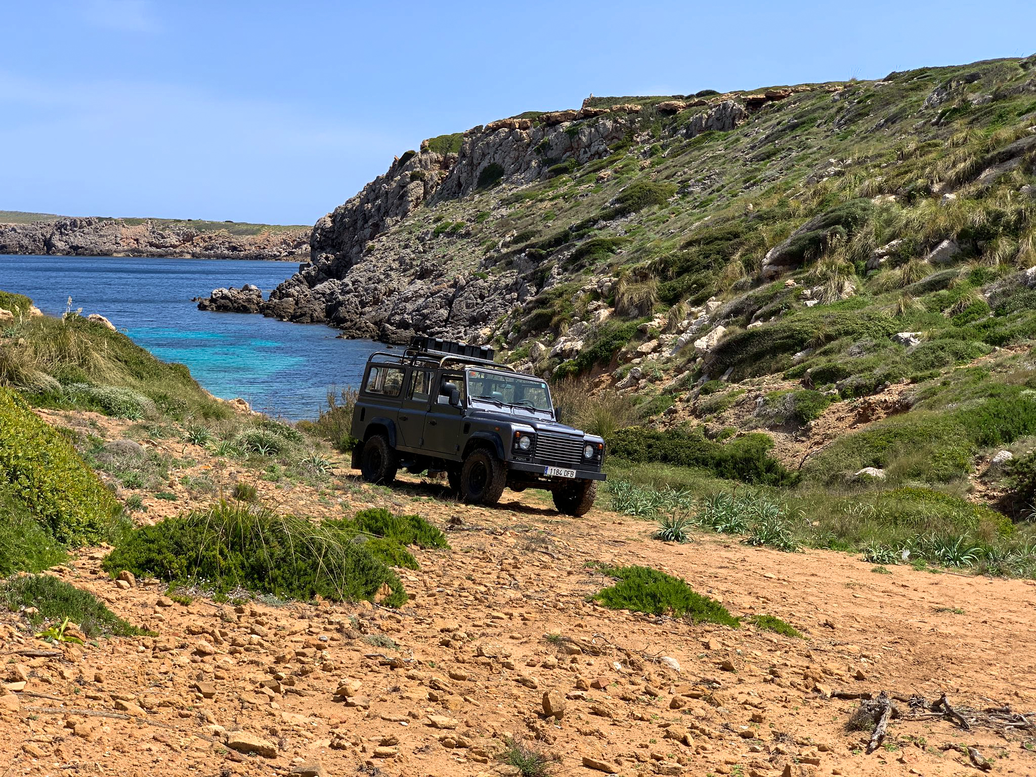 Land Rover Defender en sendero costero, mar turquesa y acantilados rocosos.