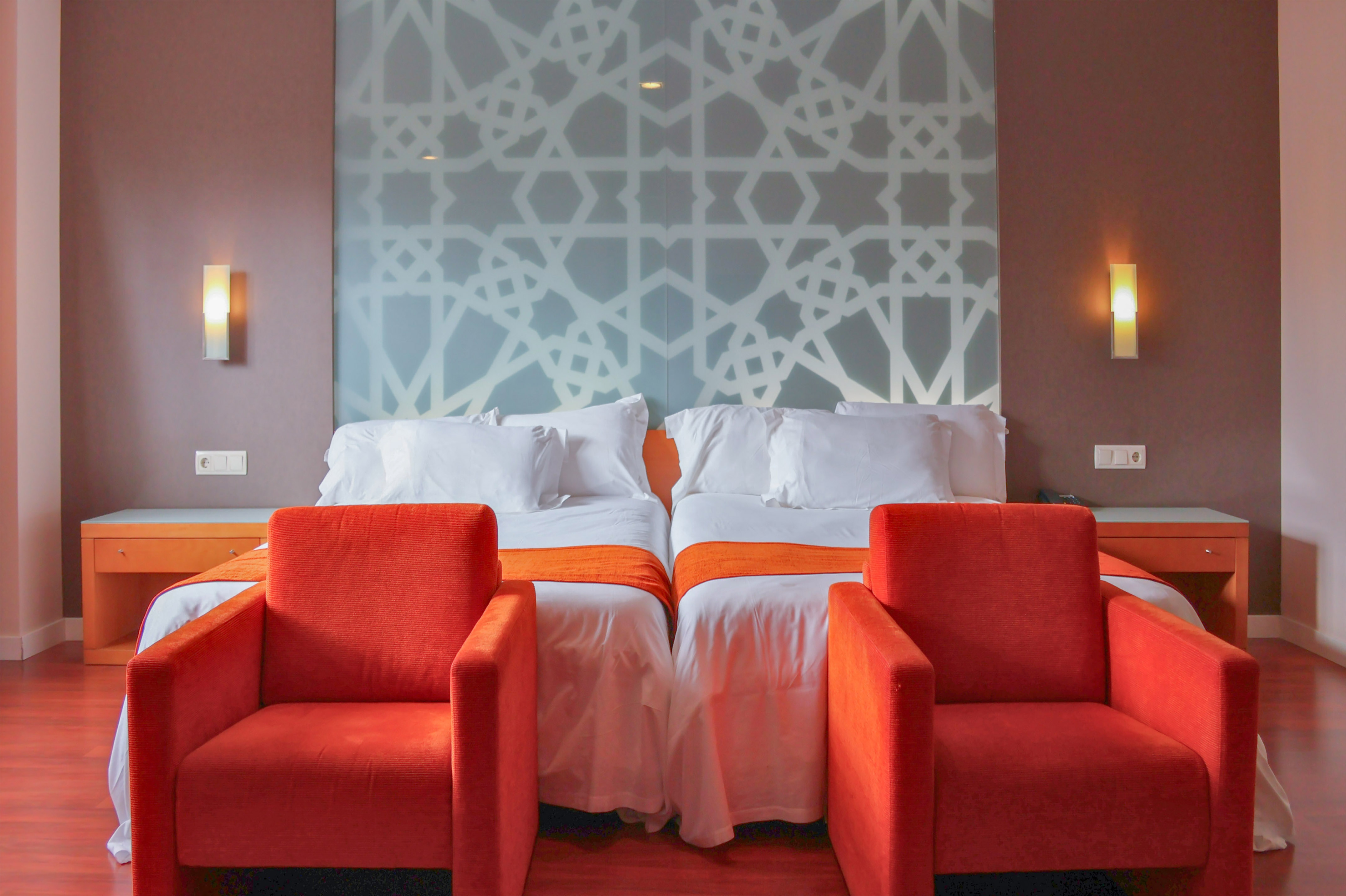 Hotel room with twin beds, orange armchairs, and a geometric pattern headboard.