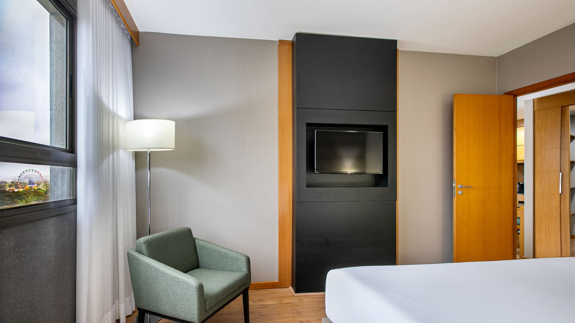 Hotel room with bed, armchair, TV, and window view of a Ferris wheel.