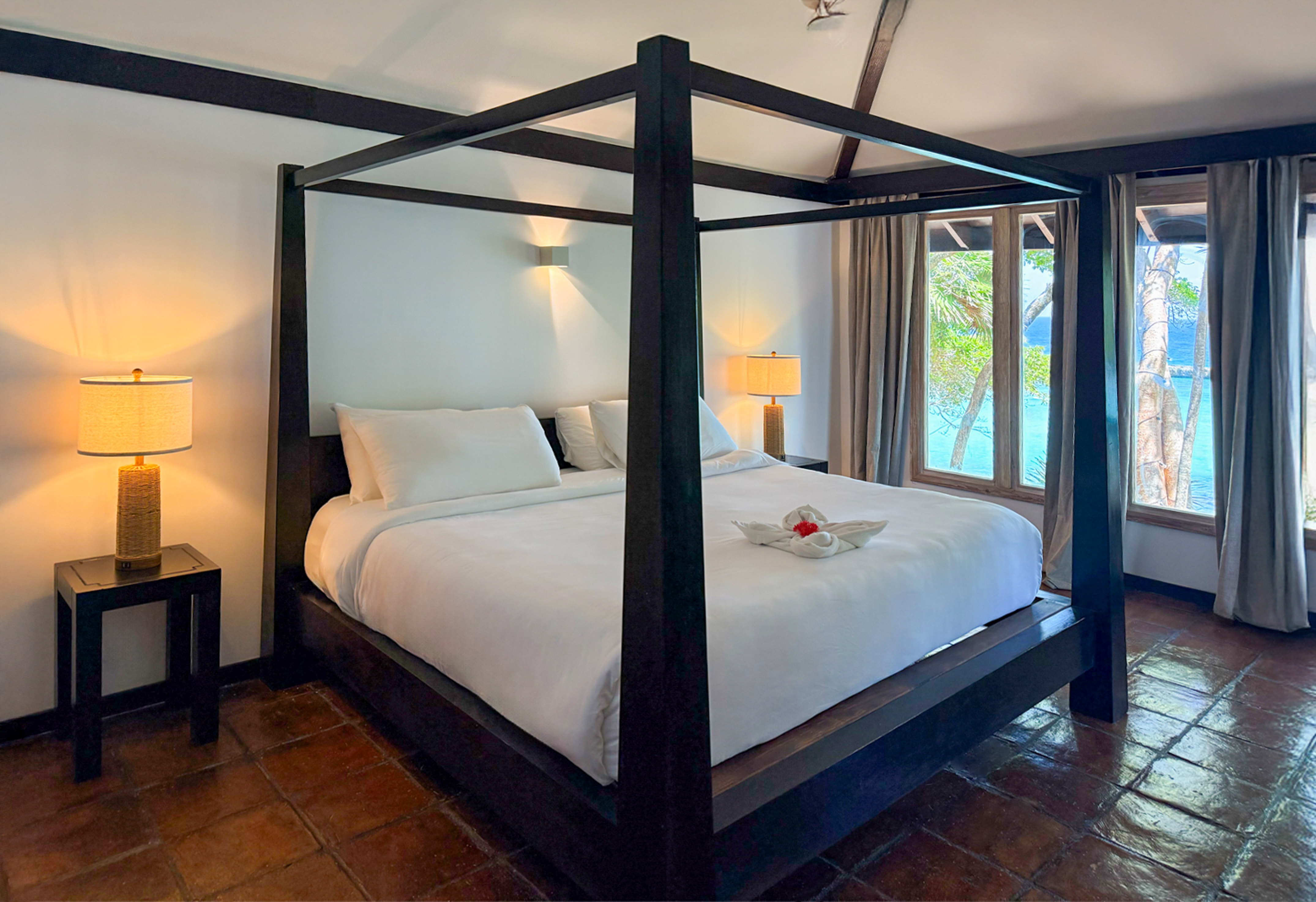 Tropical bedroom with dark four-poster bed, white linens, ocean view.
