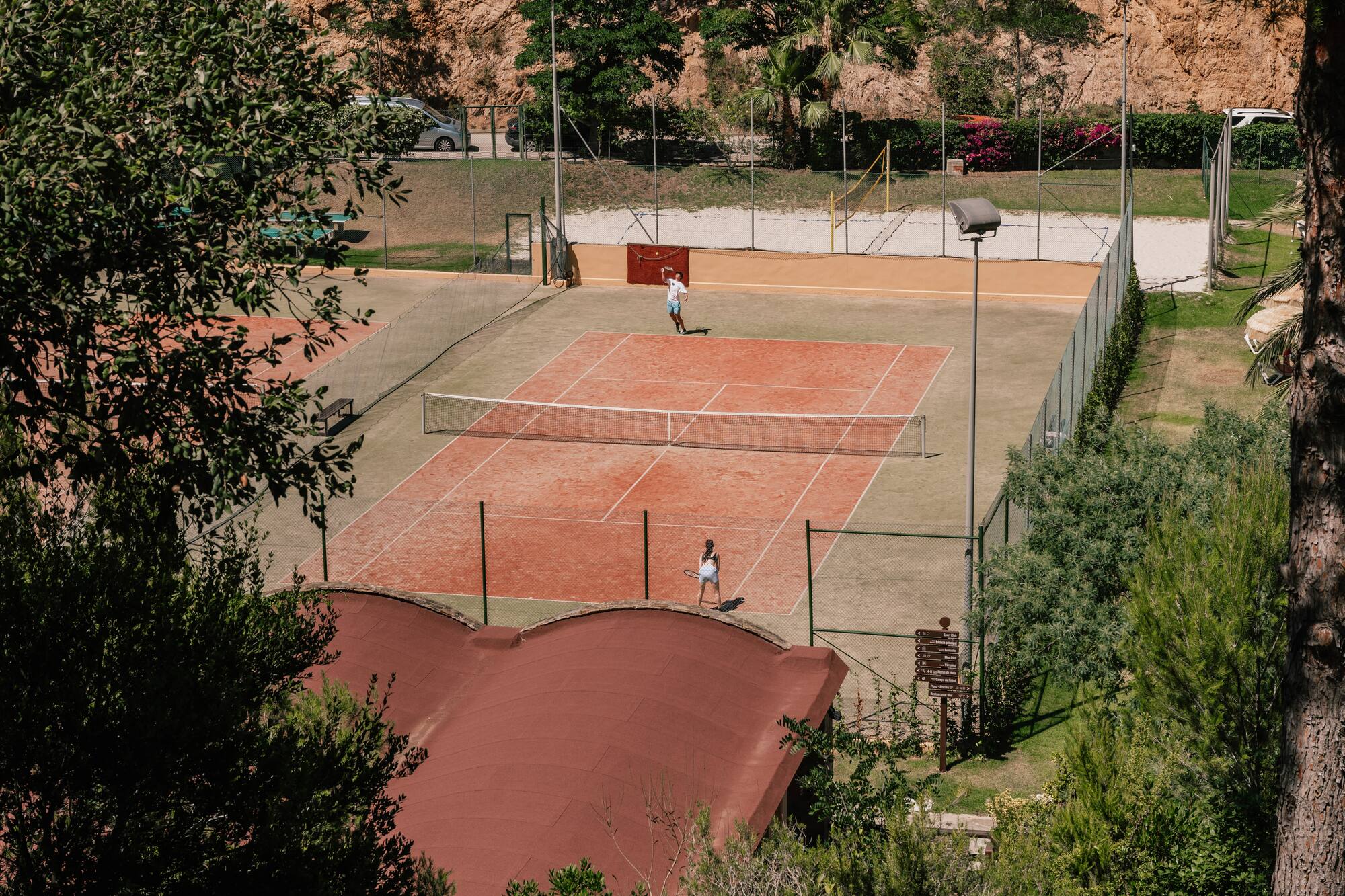 Players on a red clay tennis court, surrounded by lush green hills and trees.