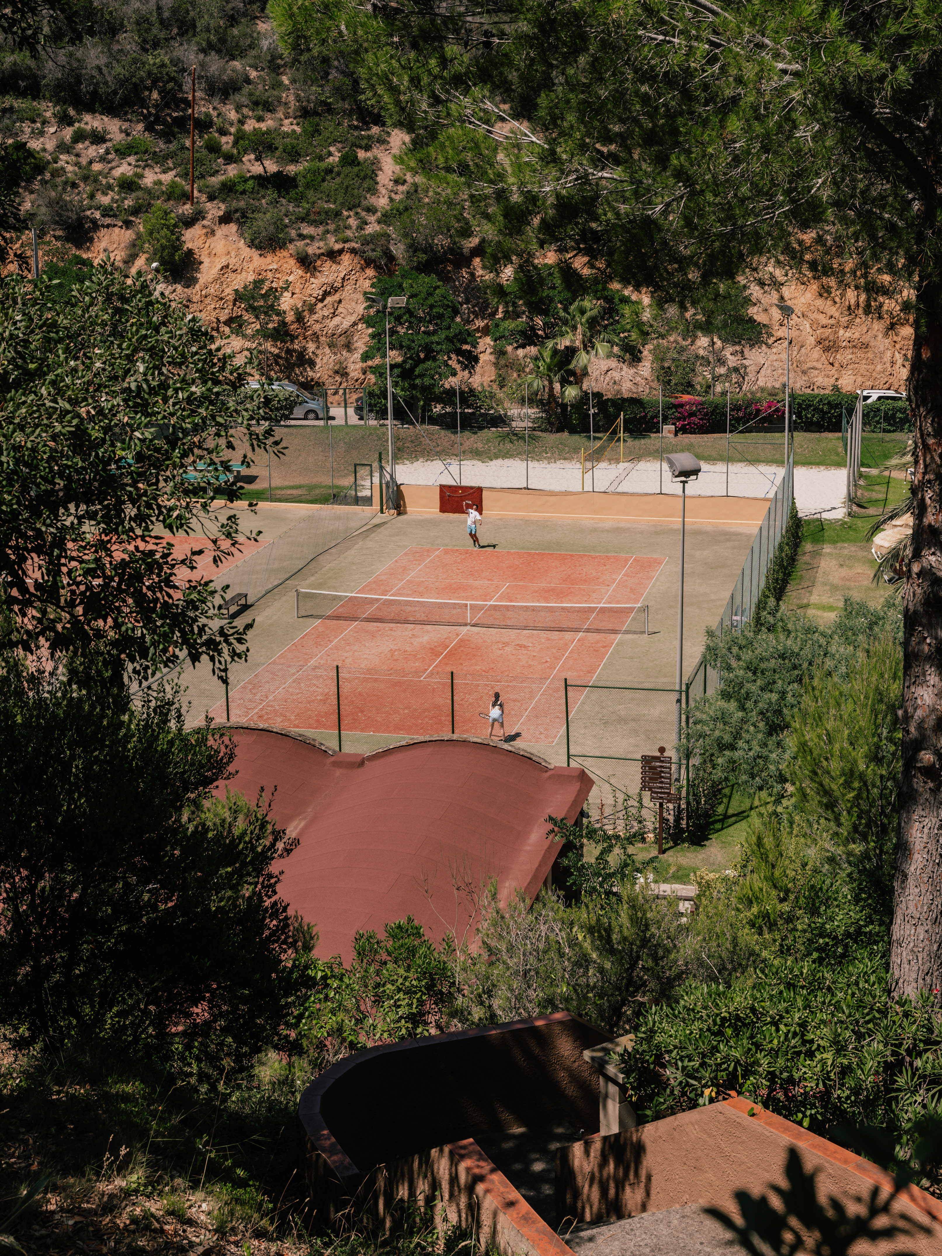Players on a red clay tennis court, surrounded by lush green hills and trees.
