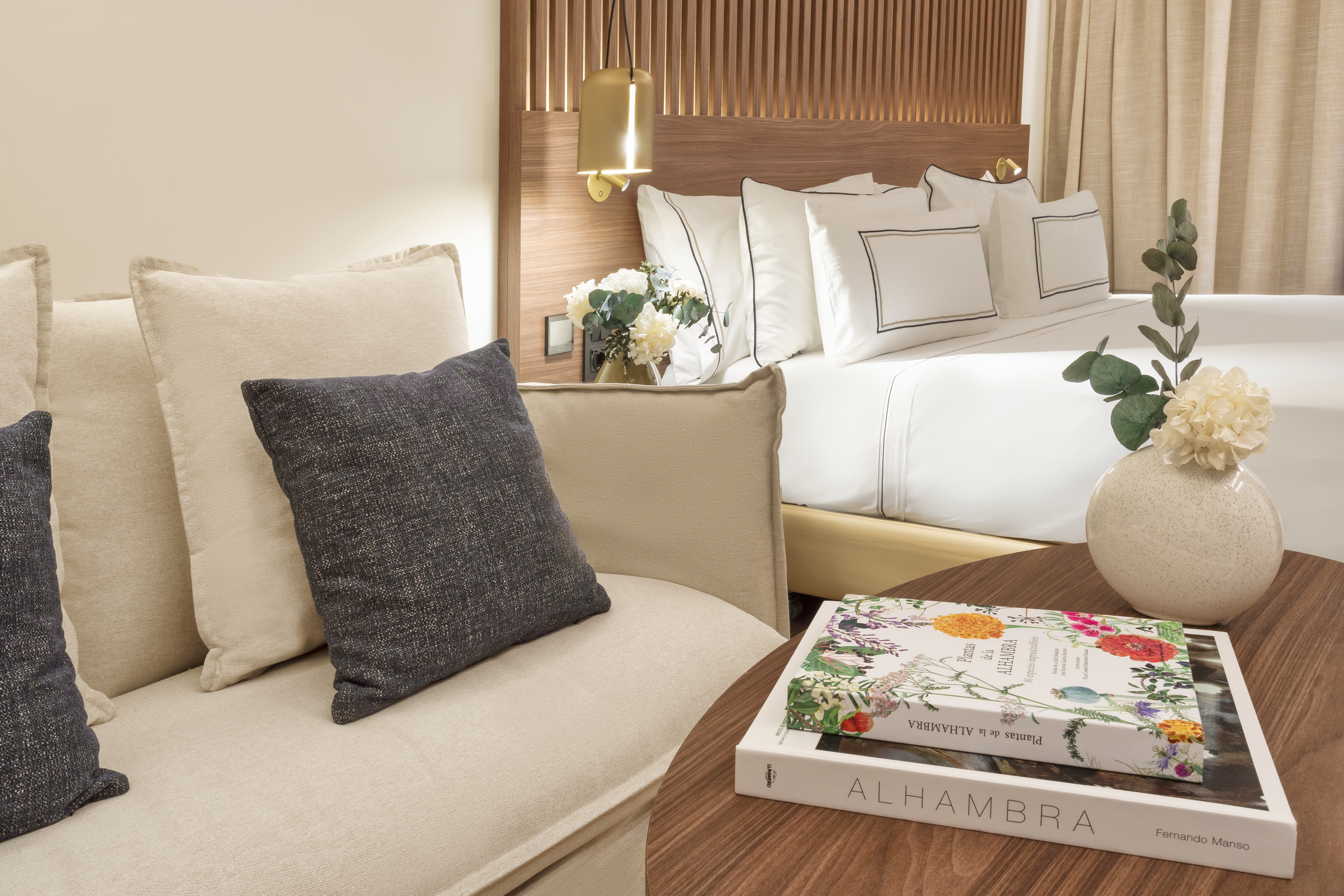 Modern hotel bedroom with white bed, cozy sofa, Alhambra books, and floral decor.