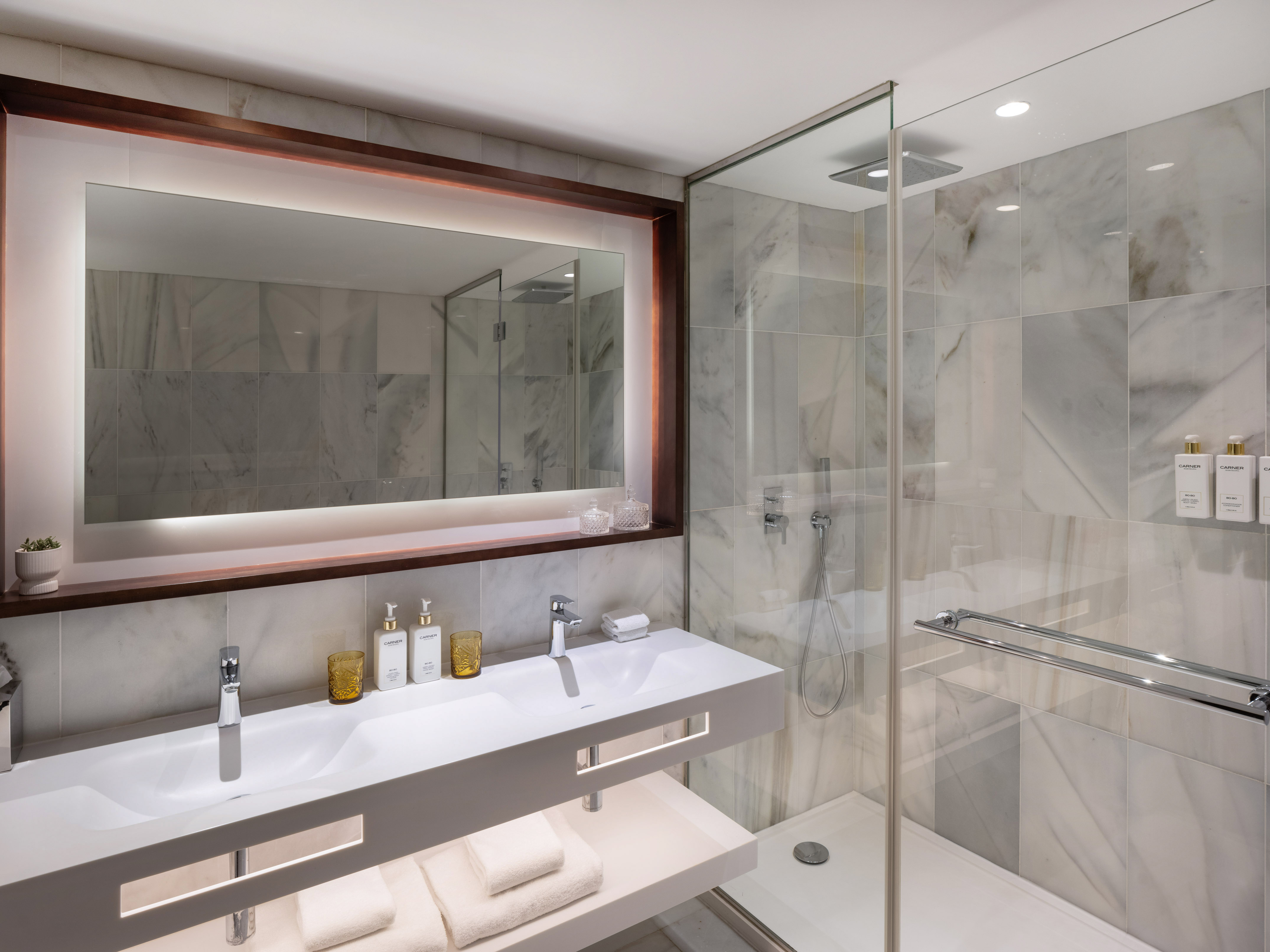 Modern hotel bathroom with double vanity, backlit mirror, and glass shower.