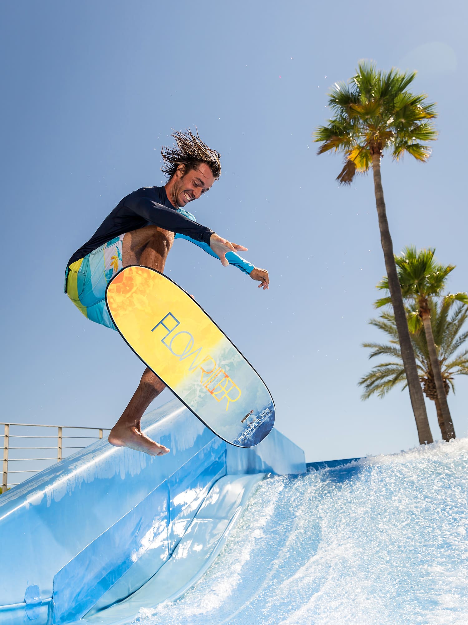 Surfer jumping on Flowrider wave simulator, palm trees, blue sky.