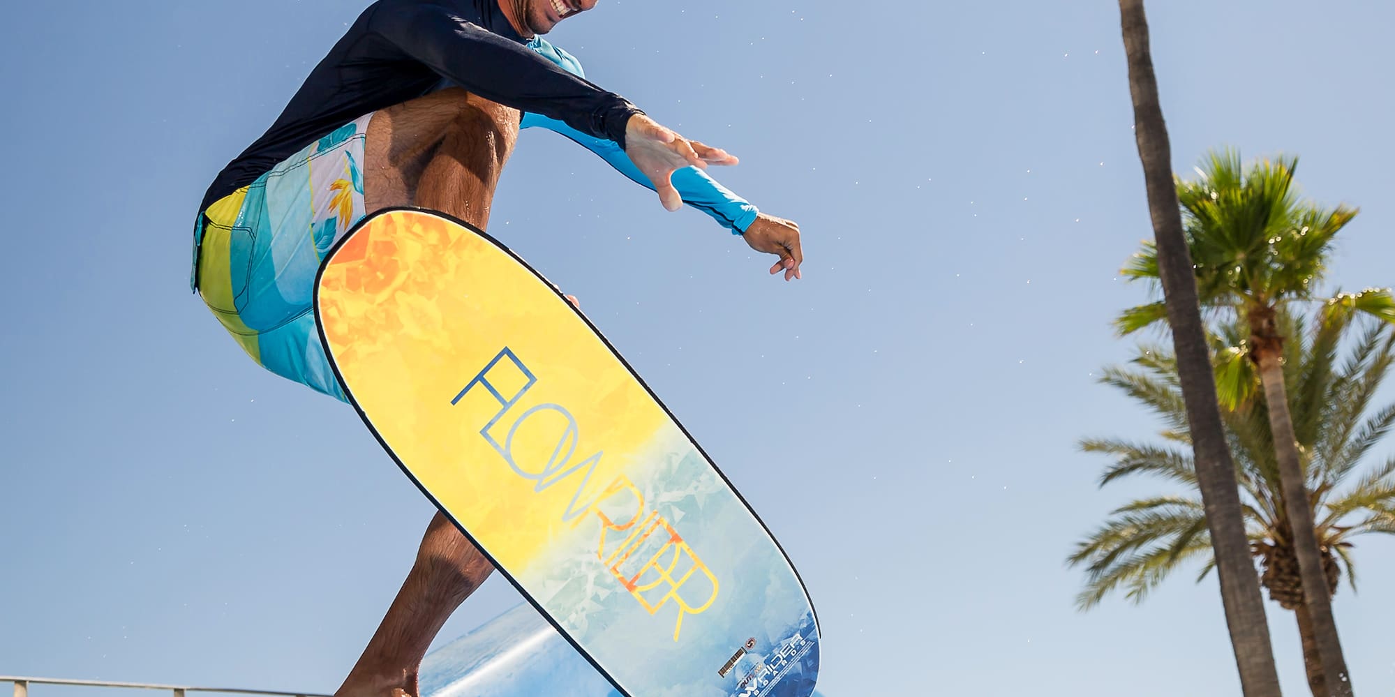 Man performs aerial jump on FlowRider surf simulator under palm trees.
